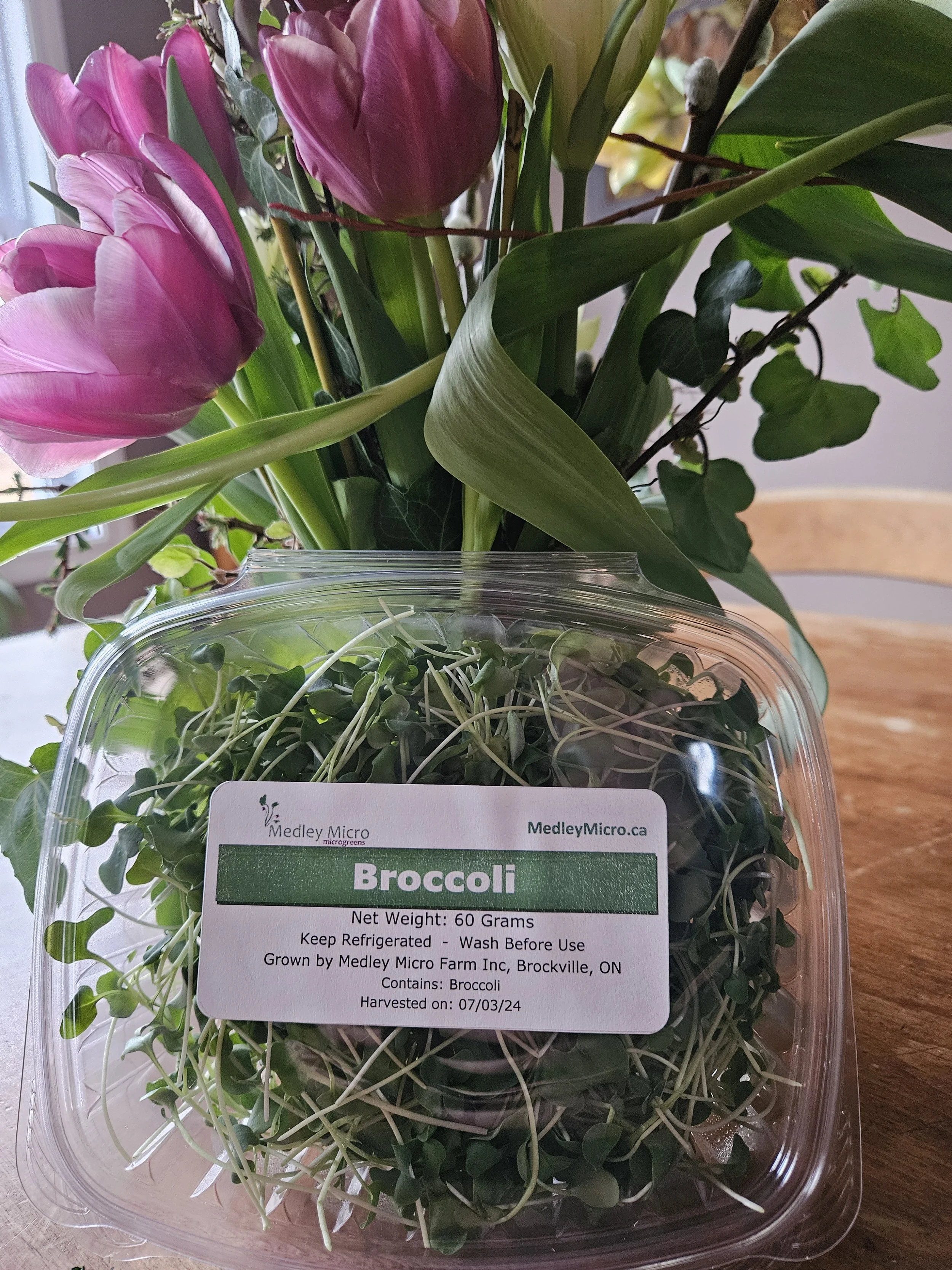 Plastic container of broccoli microgreens with label, placed next to a vase of purple and white tulips on a wooden table.
