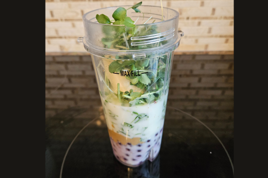 Blender cup with layered smoothie ingredients including blueberries, banana, yogurt, and greens, on a dark countertop.