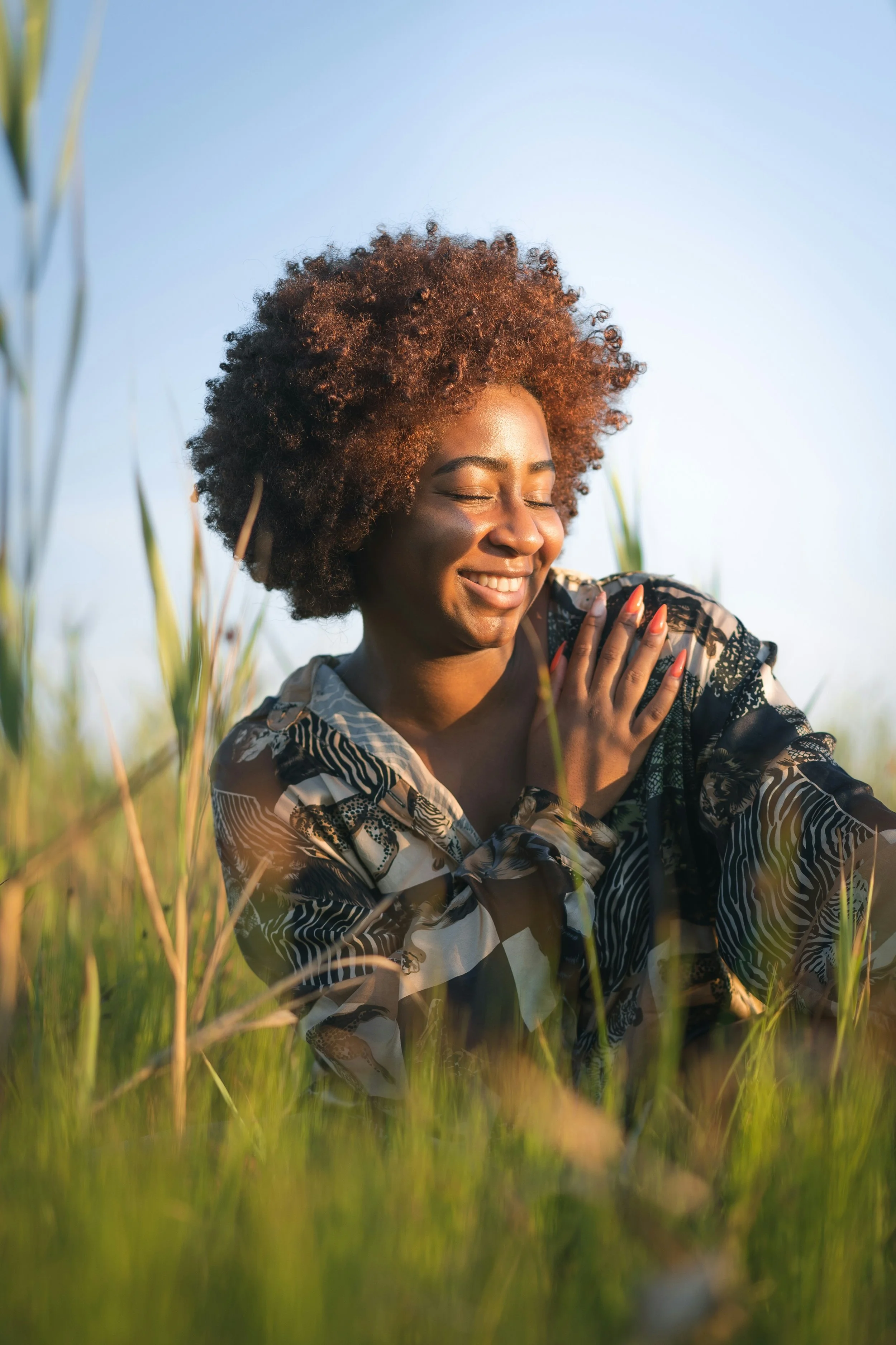 A woman with curly hair smiling with her eyes closed, standing in a field of tall grass during sunset.