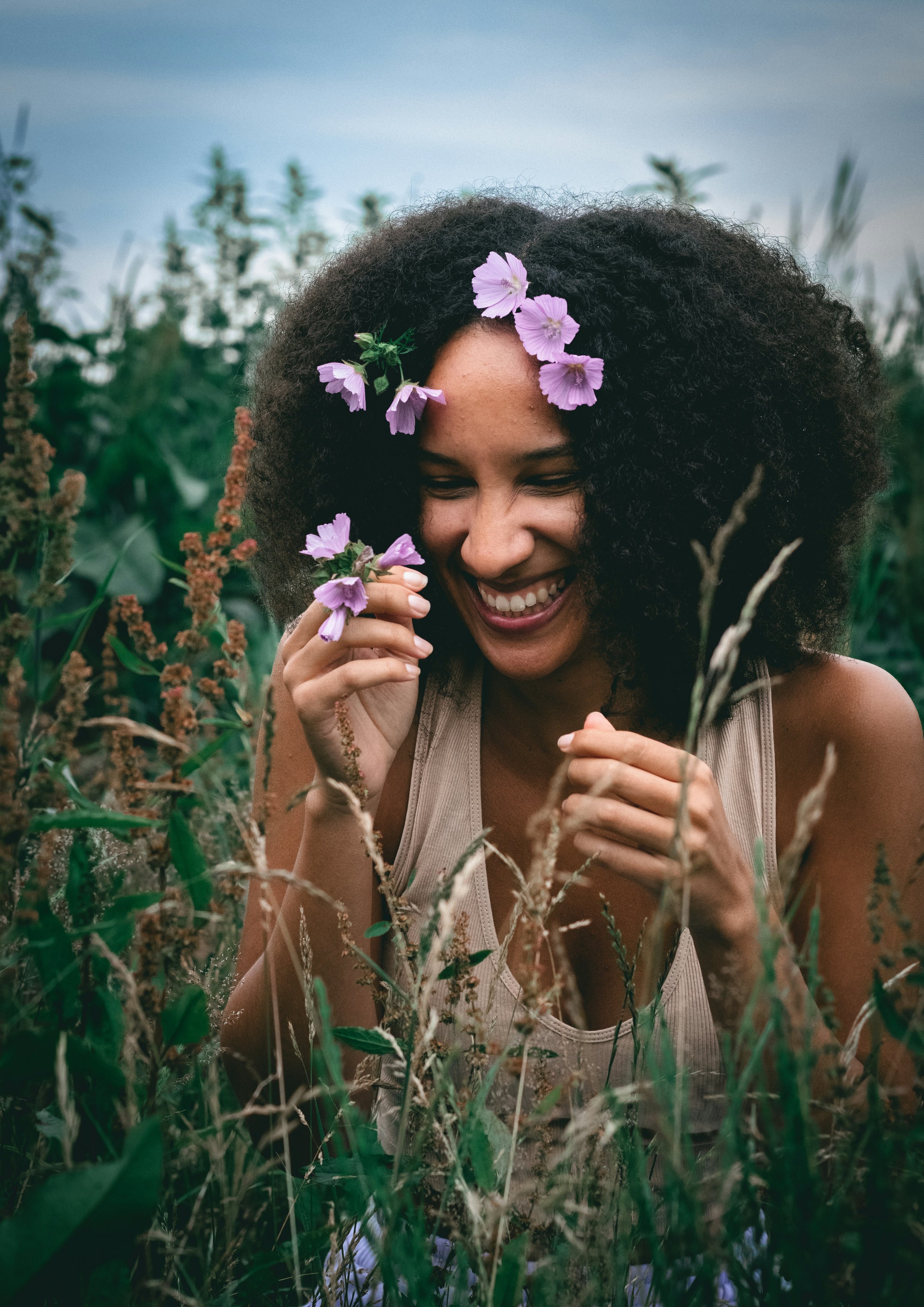 Woman with curly hair smiling and holding purple flowers in a green field during daytime.