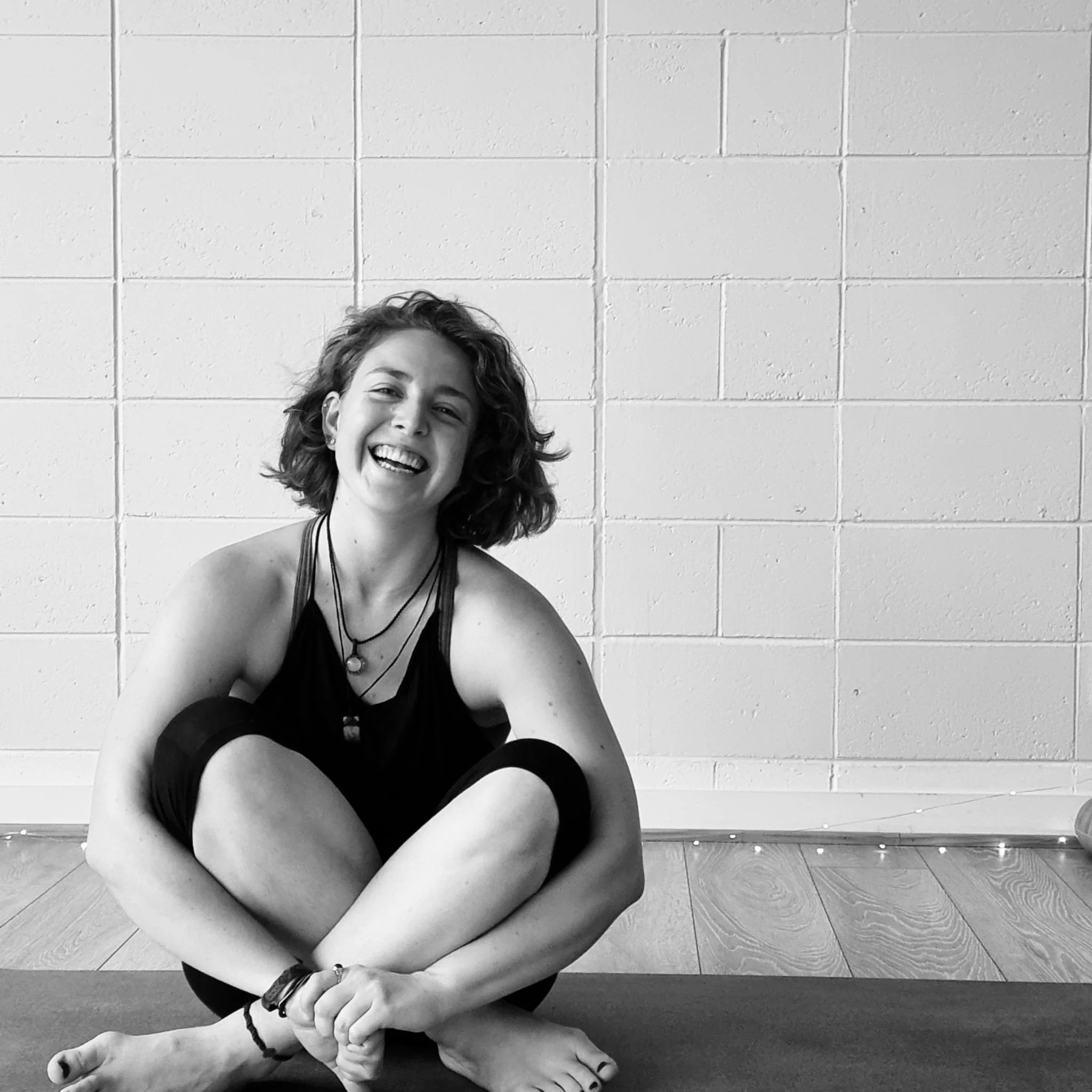 Young woman with curly hair smiling and sitting on the floor in front of a brick wall.