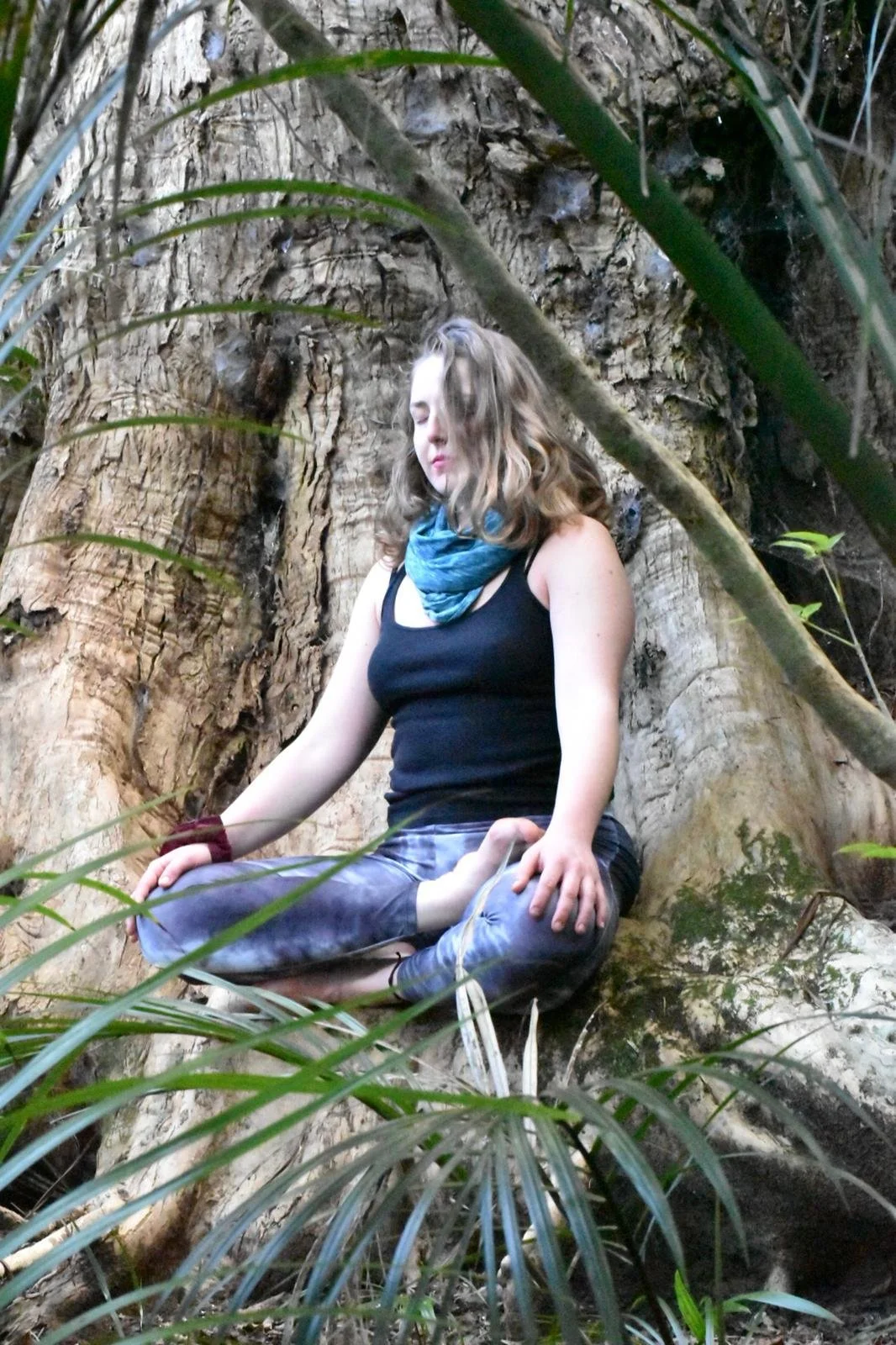 A young woman in a black tank top and colorful leggings is seated cross-legged on a tree root in a dense forest, meditating with eyes closed and a blue scarf around her neck.