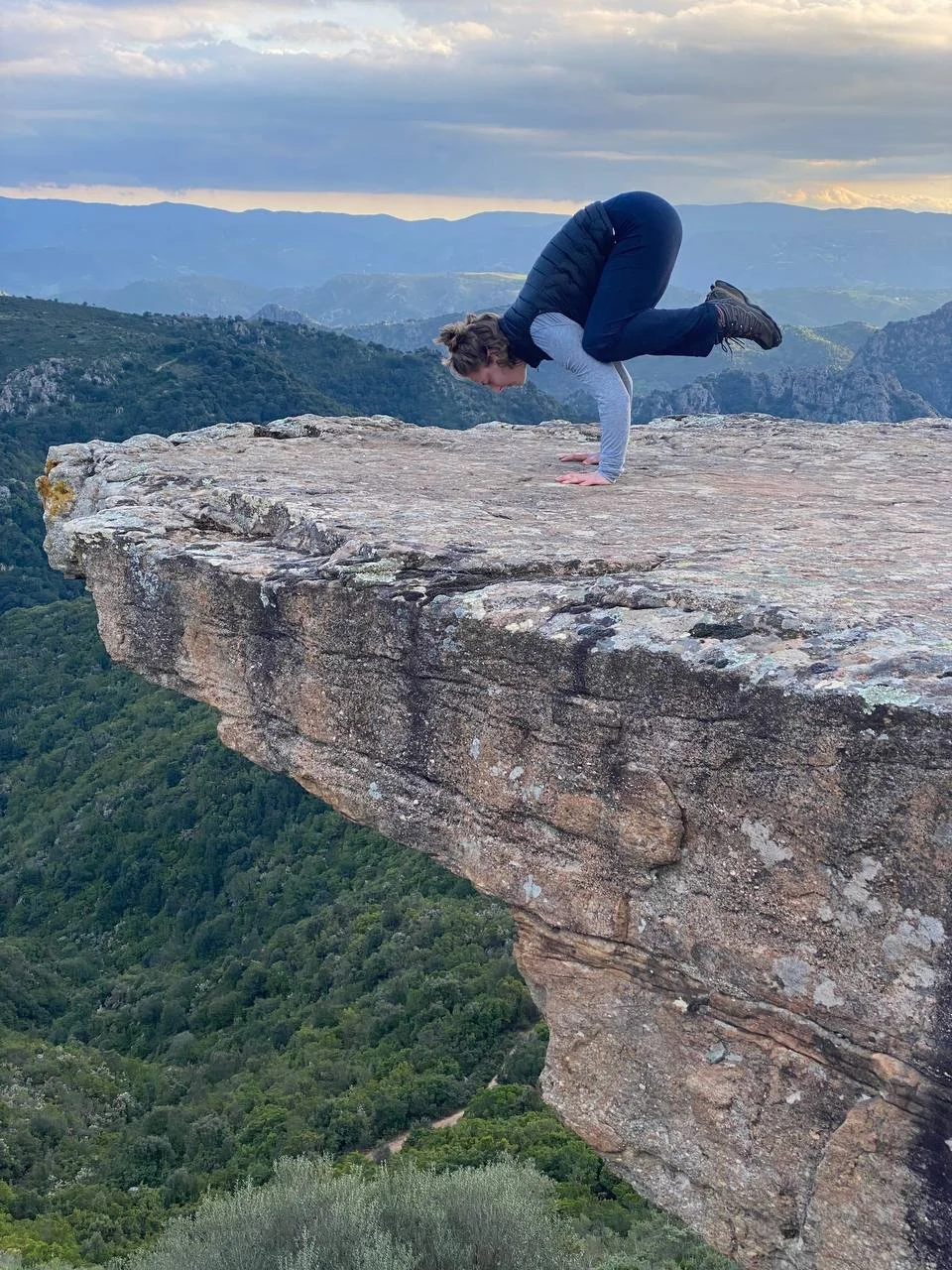 A person performing a handstand on the edge of a large mountain ledge with a valley and distant mountains in the background.