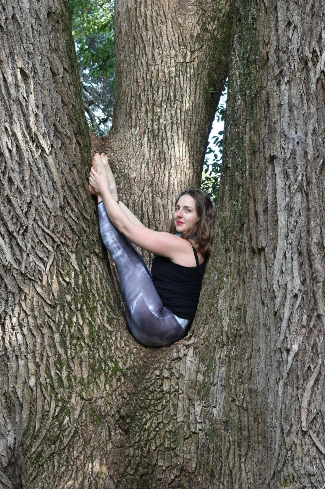 A woman sitting in the fork of a large tree, holding her feet with both hands, wearing black tank top and patterned leggings, with a slight smile.
