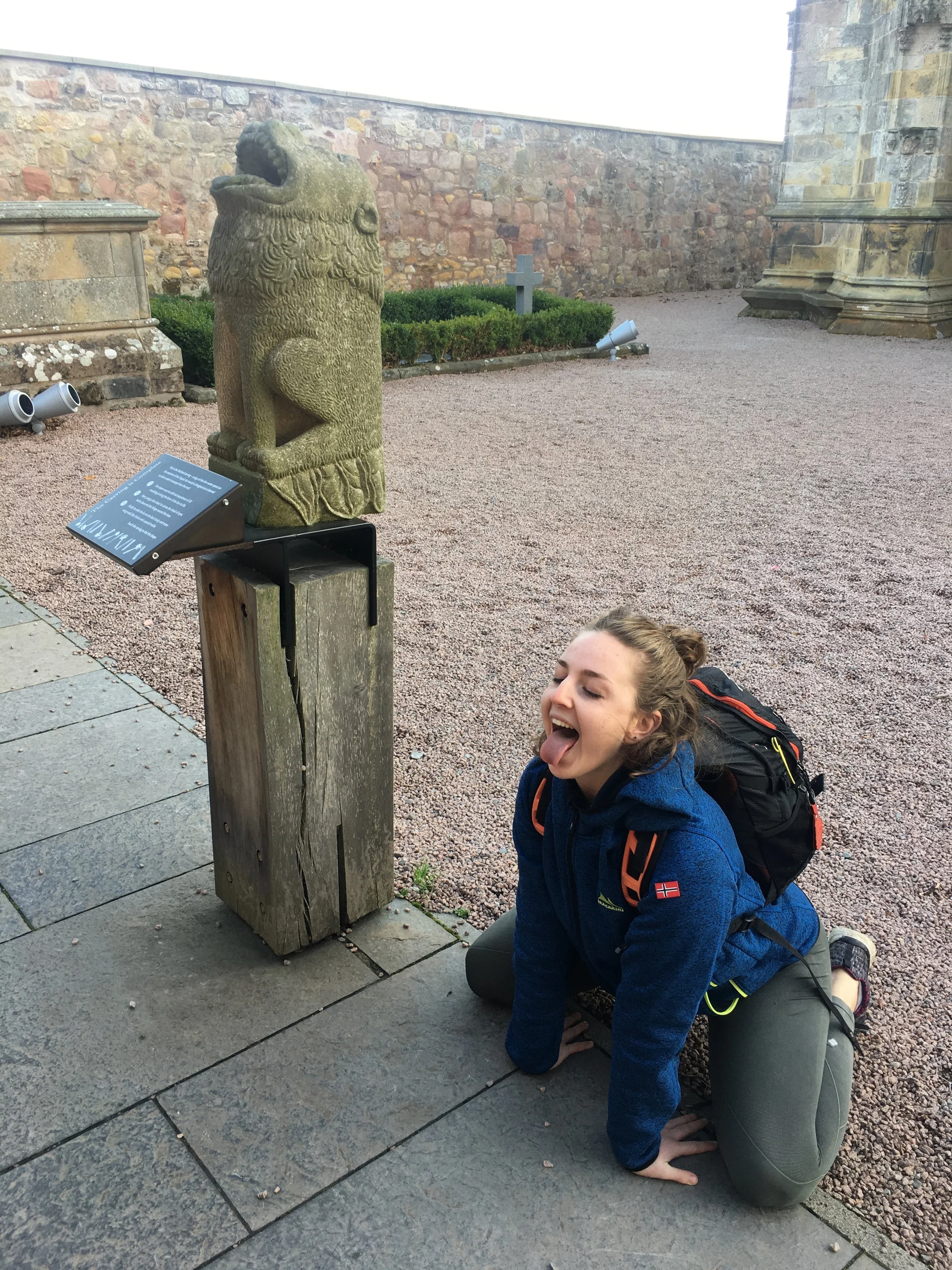 A young woman with a backpack in a crouching position on the ground making a funny face with her tongue out, next to a stone lion sculpture in an outdoor historical site with stone walls and informational plaque.