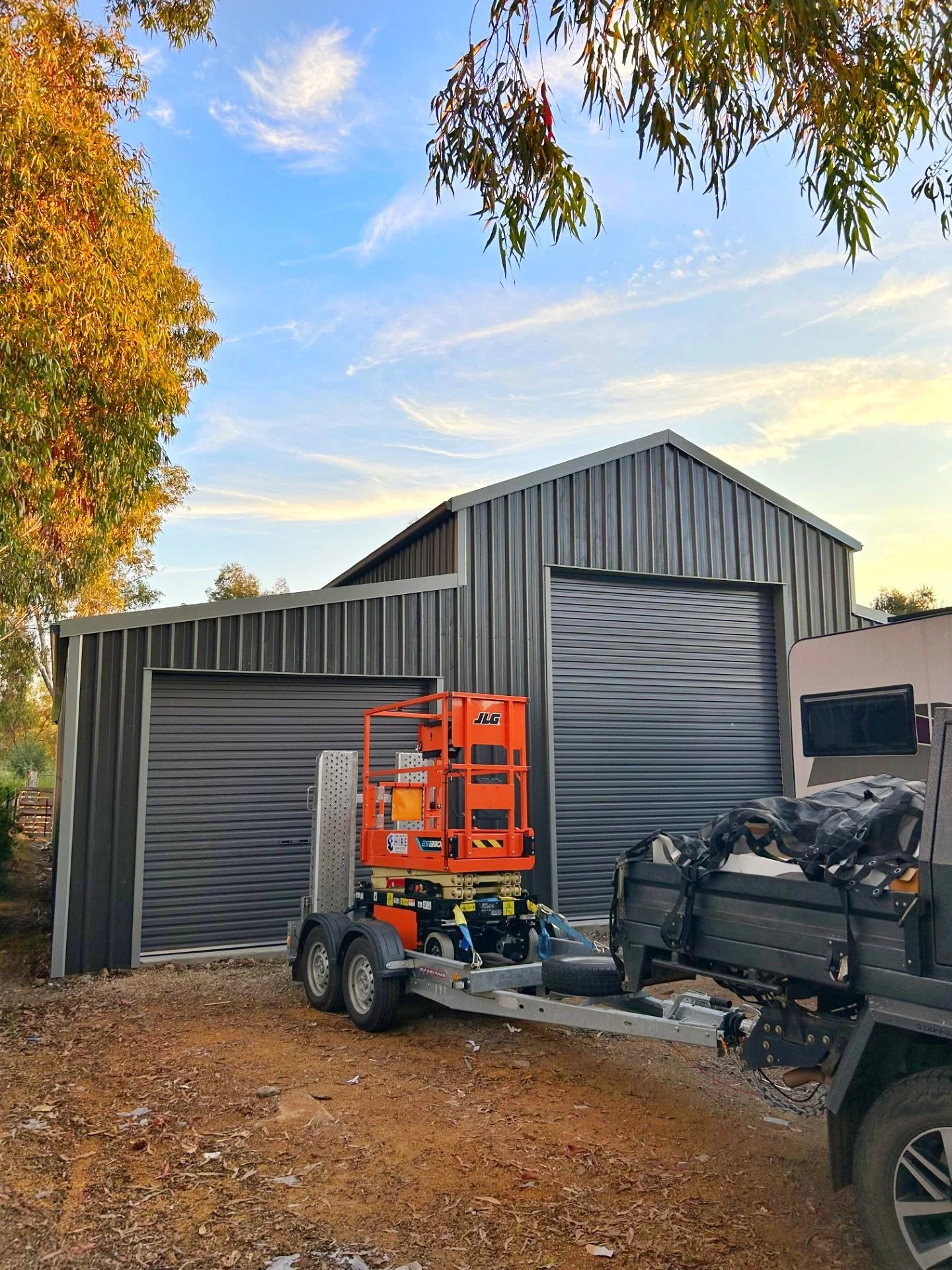 An industrial building with metal siding and a large roll-up door, outside a trailer with construction equipment, on a dirt ground with fallen leaves, under a partly cloudy sky with trees in the background.