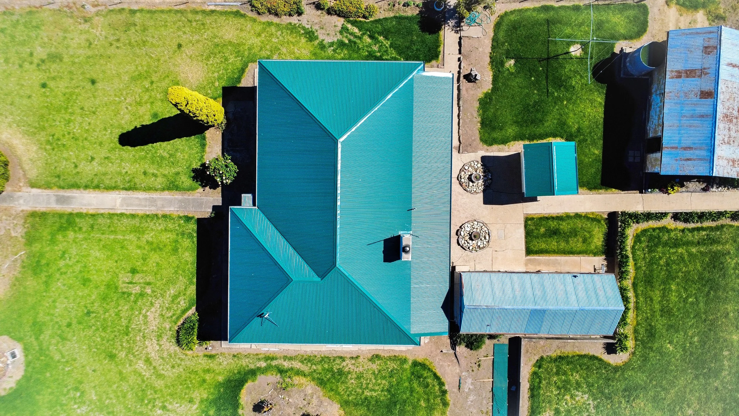 An aerial view of a house with a teal metal roof, surrounded by a well-maintained lawn, trees, and concrete walkways.