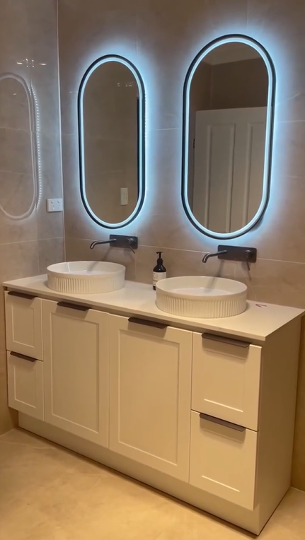Bathroom with two oval mirrors with backlit LED lighting, a white double vanity with vessel sinks, black wall-mounted faucets, a soap dispenser, and beige tiled walls.