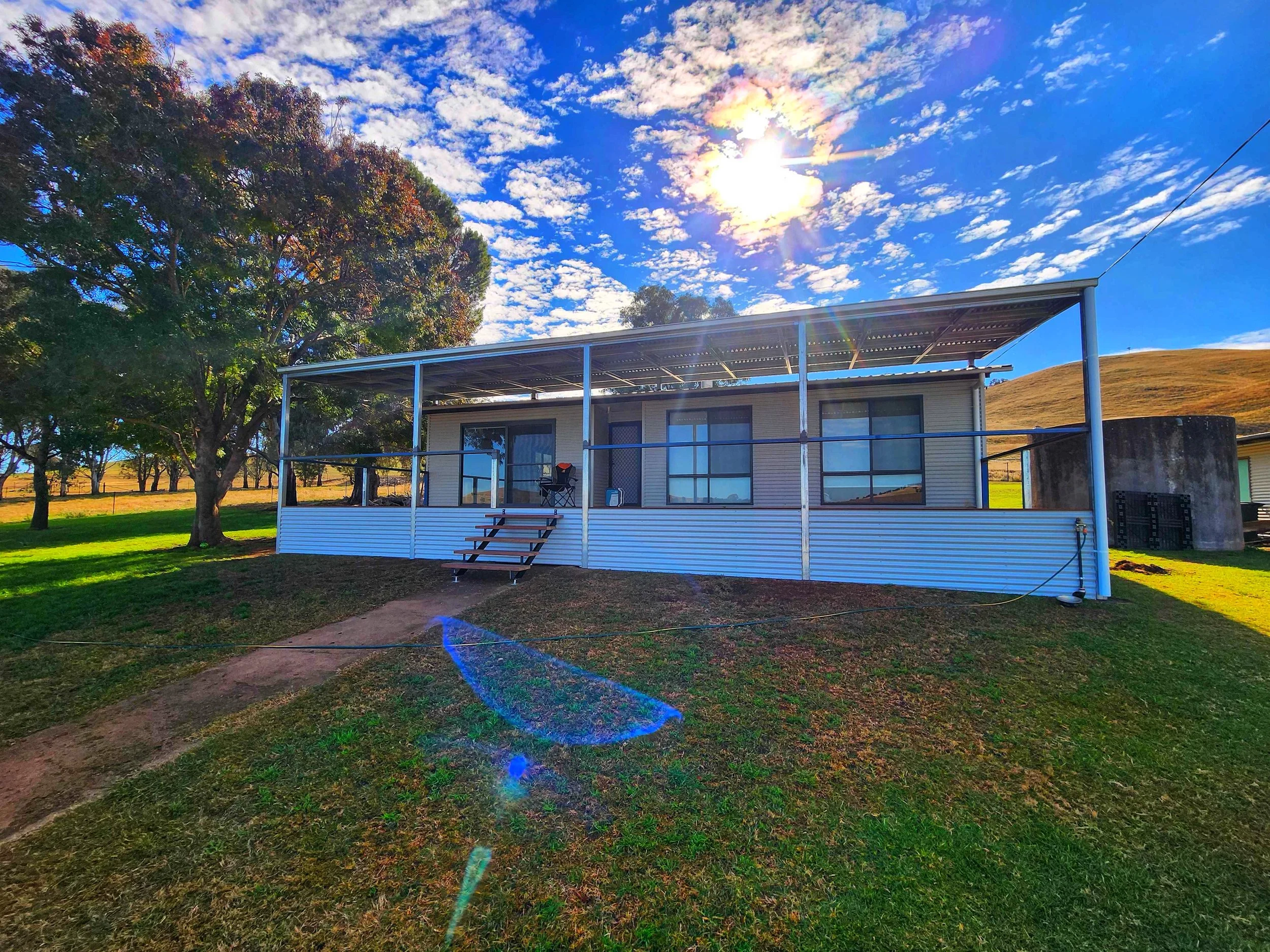 A house with a metal frame porch, large windows, and glass sliding doors is situated in a green yard with trees and a grassy hill in the background. The sky is partly cloudy with the sun shining brightly, creating lens flares in the image.