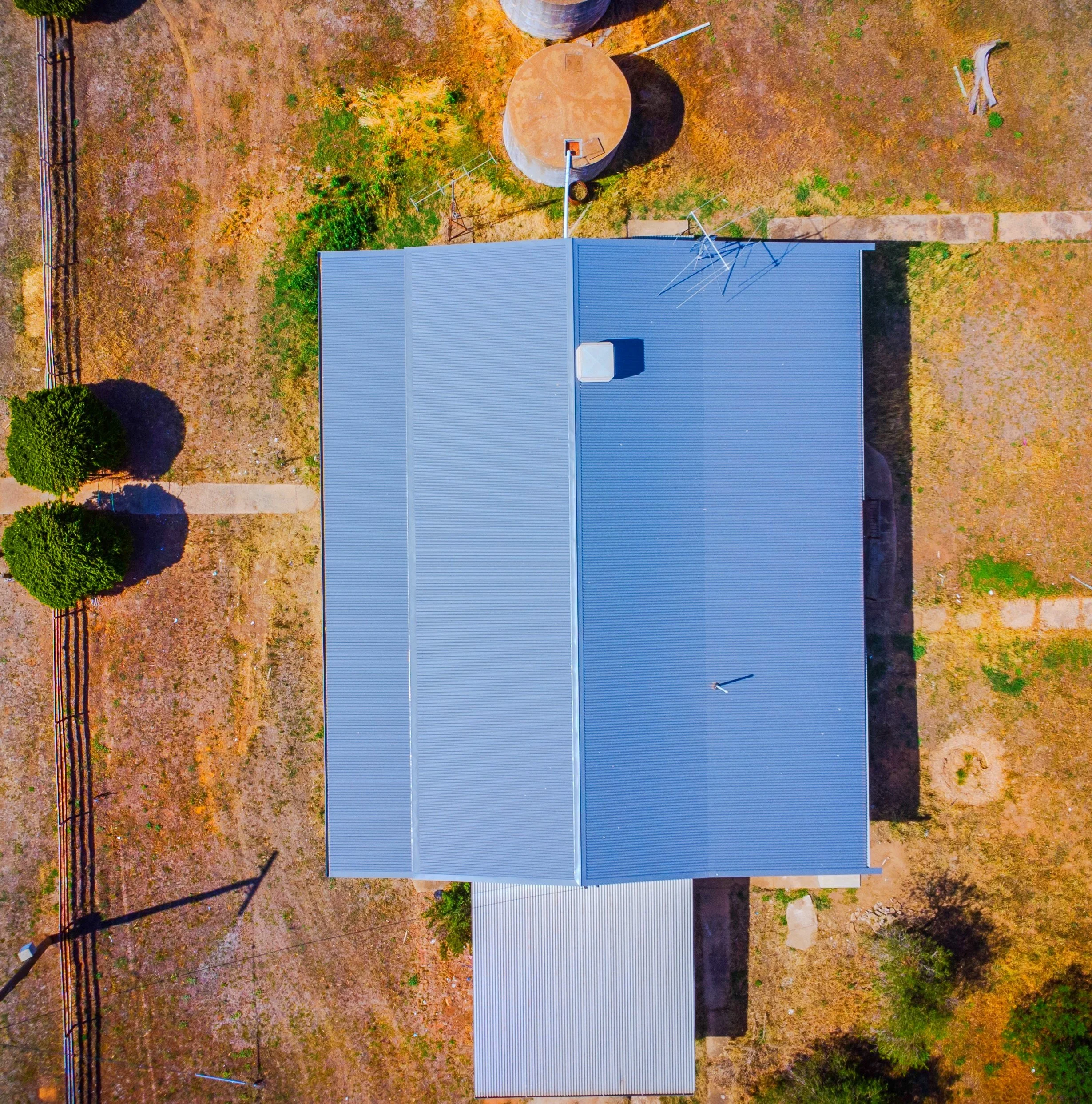 An aerial view of a blue metal building with a pitched roof, several antennas or satellite dishes attached, and a small white structure on the roof. Surrounding the building is a dry, brown landscape with green bushes, a few trees, a dirt sidewalk, a