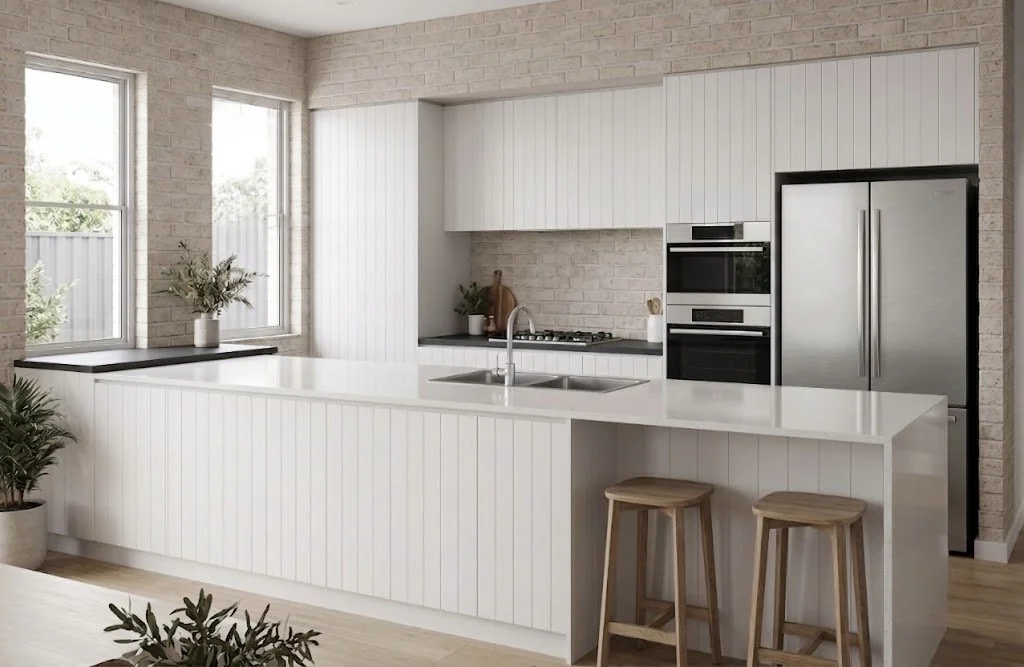 Modern white kitchen with brick walls, large windows, a central island with a sink, and stainless steel appliances including a refrigerator and built-in oven. Two wooden stools are placed at the island.