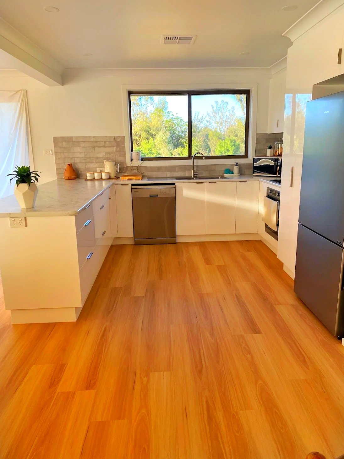 A modern kitchen with white cabinets, a gray countertop, and a large window overlooking greenery. The floor is wooden, and there are various kitchen appliances and decorative items on the counters.