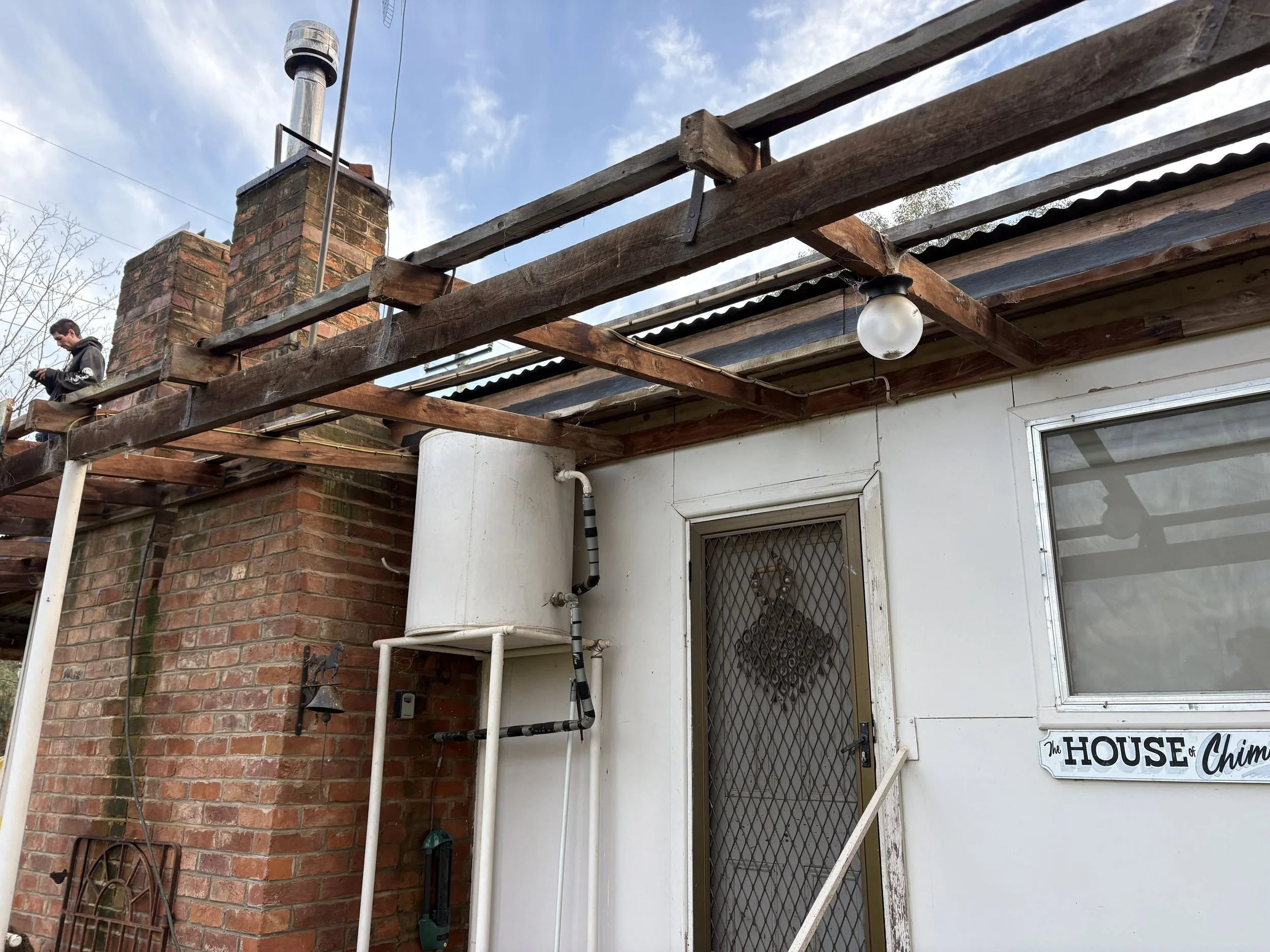 View of a house exterior with a brick chimney, wooden beams, and an outdoor light fixture. There is a person working on the roof, and a sign that reads 'The House of Chimes' is visible near the door.