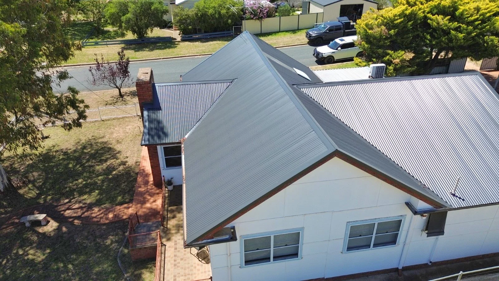 An aerial view of a house with a metal roof and white exterior walls, surrounded by a yard, trees, and a street with parked cars.