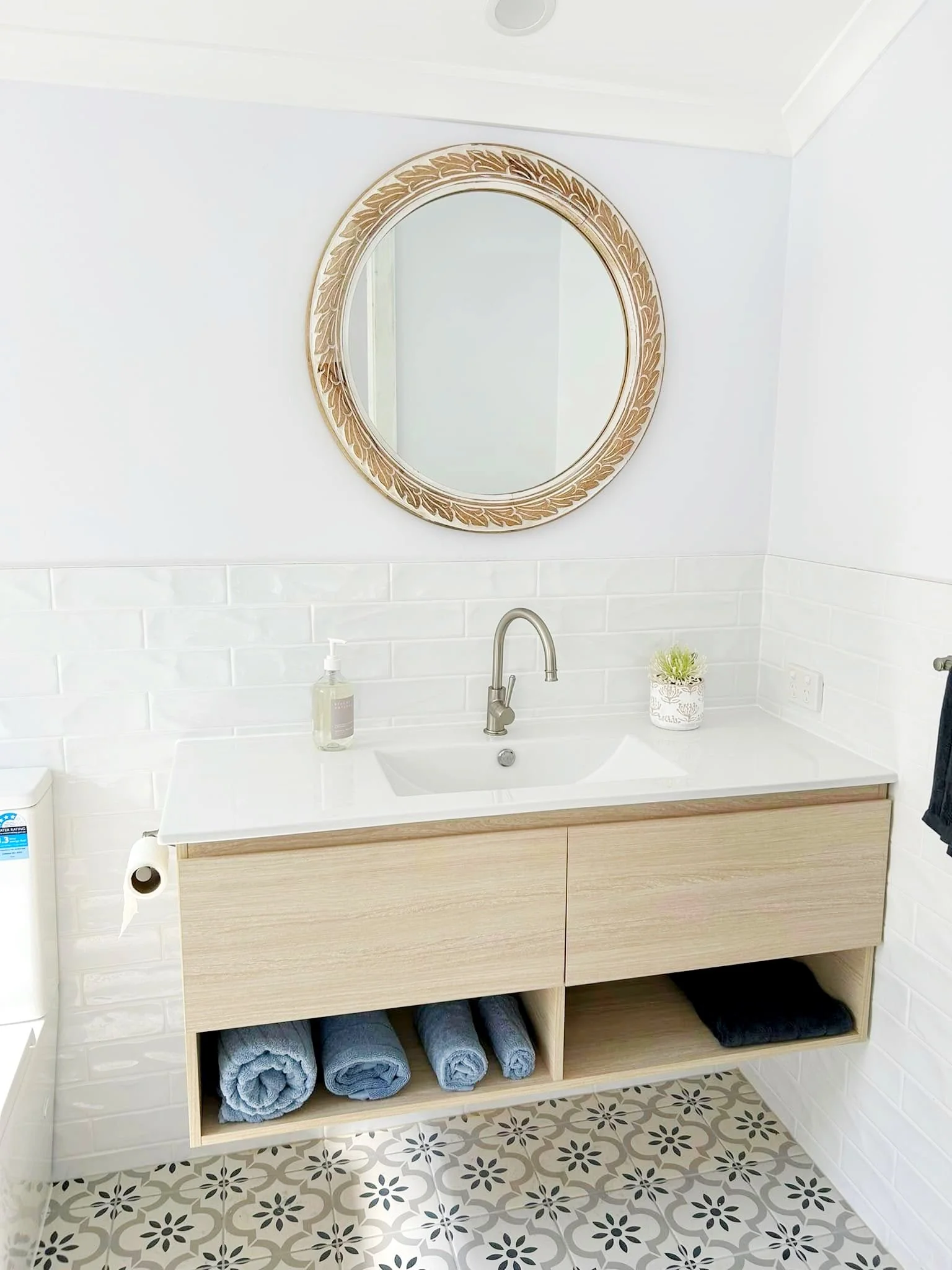 Bathroom vanity with a mirror above a wooden cabinet, towels stored in cubbies, a soap dispenser, a soap pump, a container with a plant, and a patterned floor.