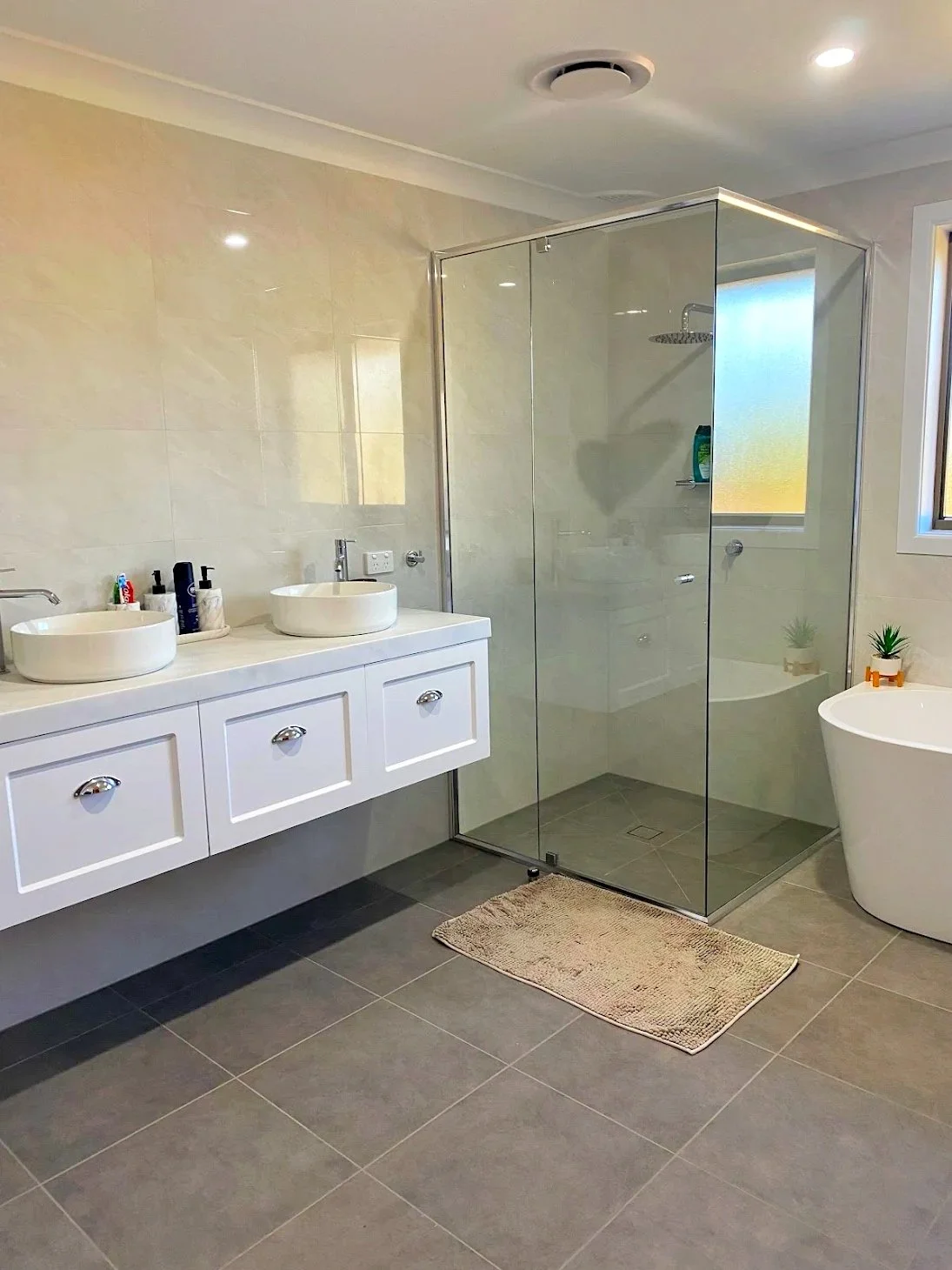 A modern bathroom featuring a glass shower enclosure, double sinks on a white vanity with storage drawers, a freestanding bathtub near a window, and small plants on a ledge. The bathroom has beige tiled walls and large dark gray floor tiles.