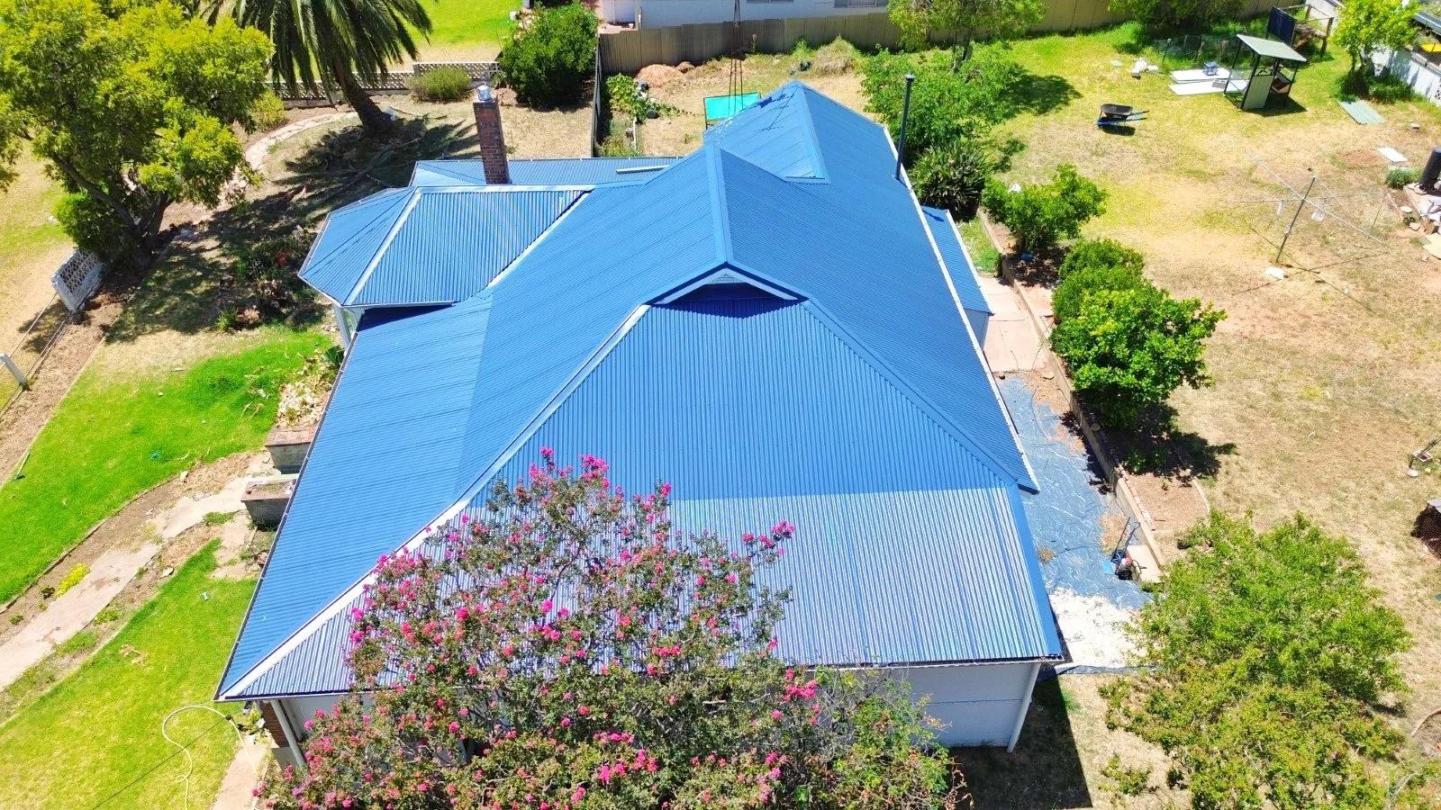 Aerial view of a house with a blue metal roof in a backyard with trees, grass, and garden areas.