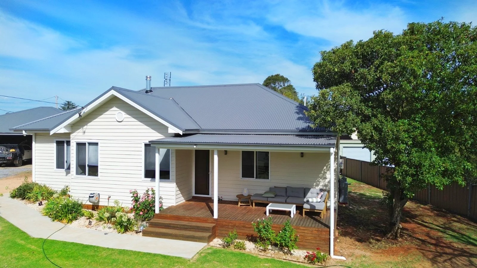 A suburban house with a grey metal roof, cream siding, a small porch with outdoor furniture, a garden with plants and flowers, a large tree, and a wooden fence under a blue sky.