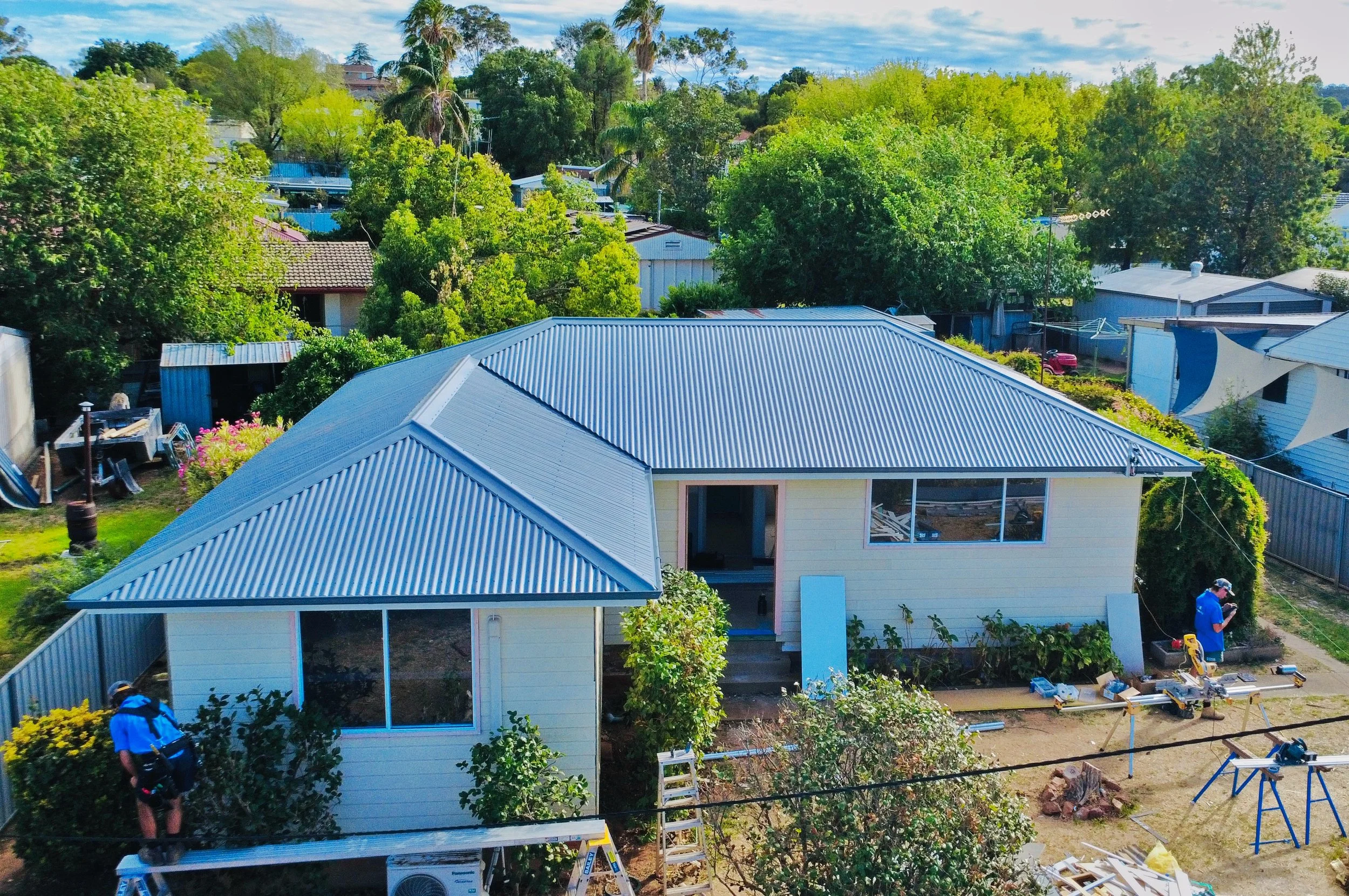 An aerial view of a house with a blue metal roof in a backyard that is under construction, with construction materials and debris scattered around.