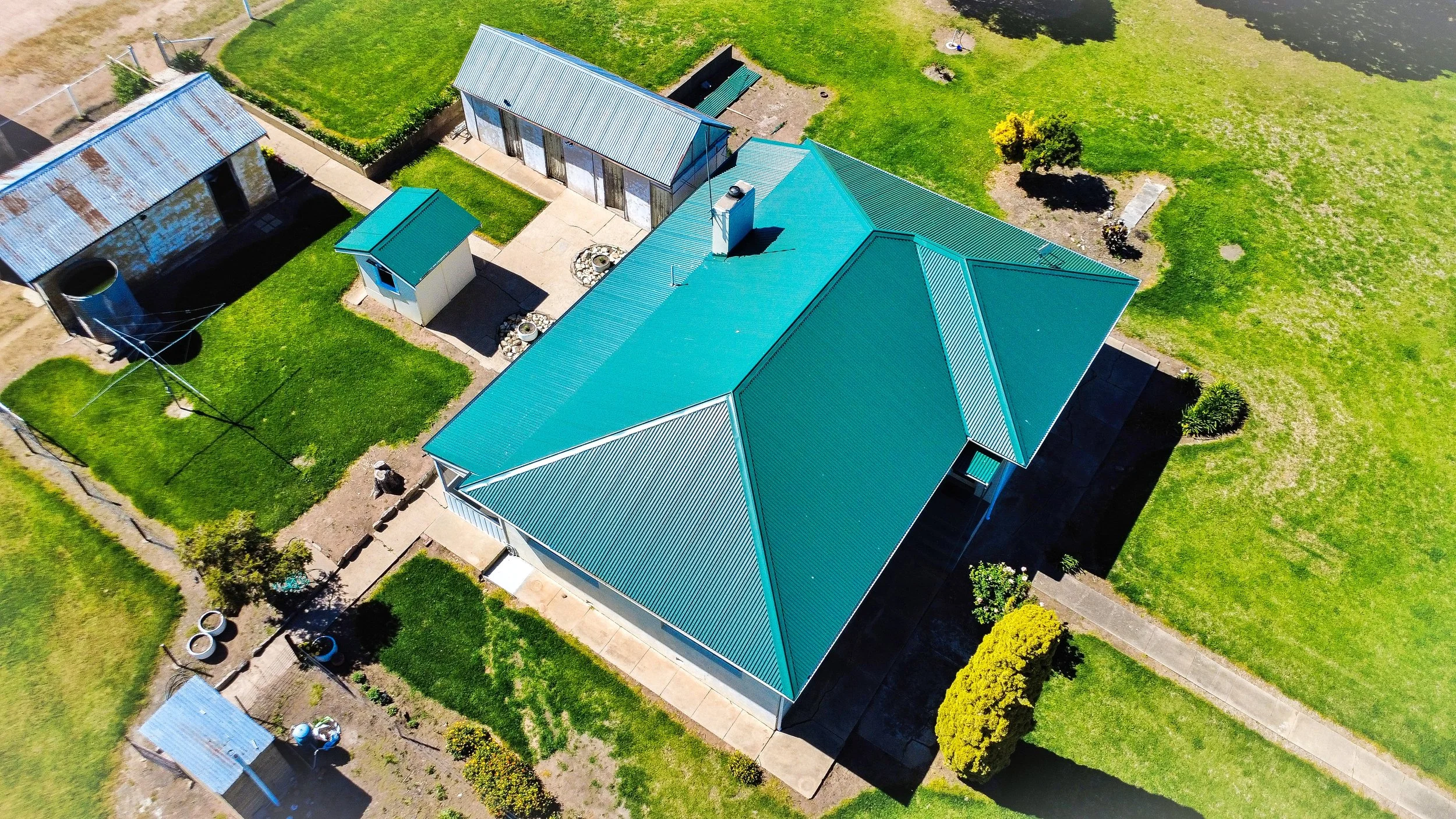 An aerial view of a house with a green metal roof, surrounded by a green lawn, pathways, and small garden areas.