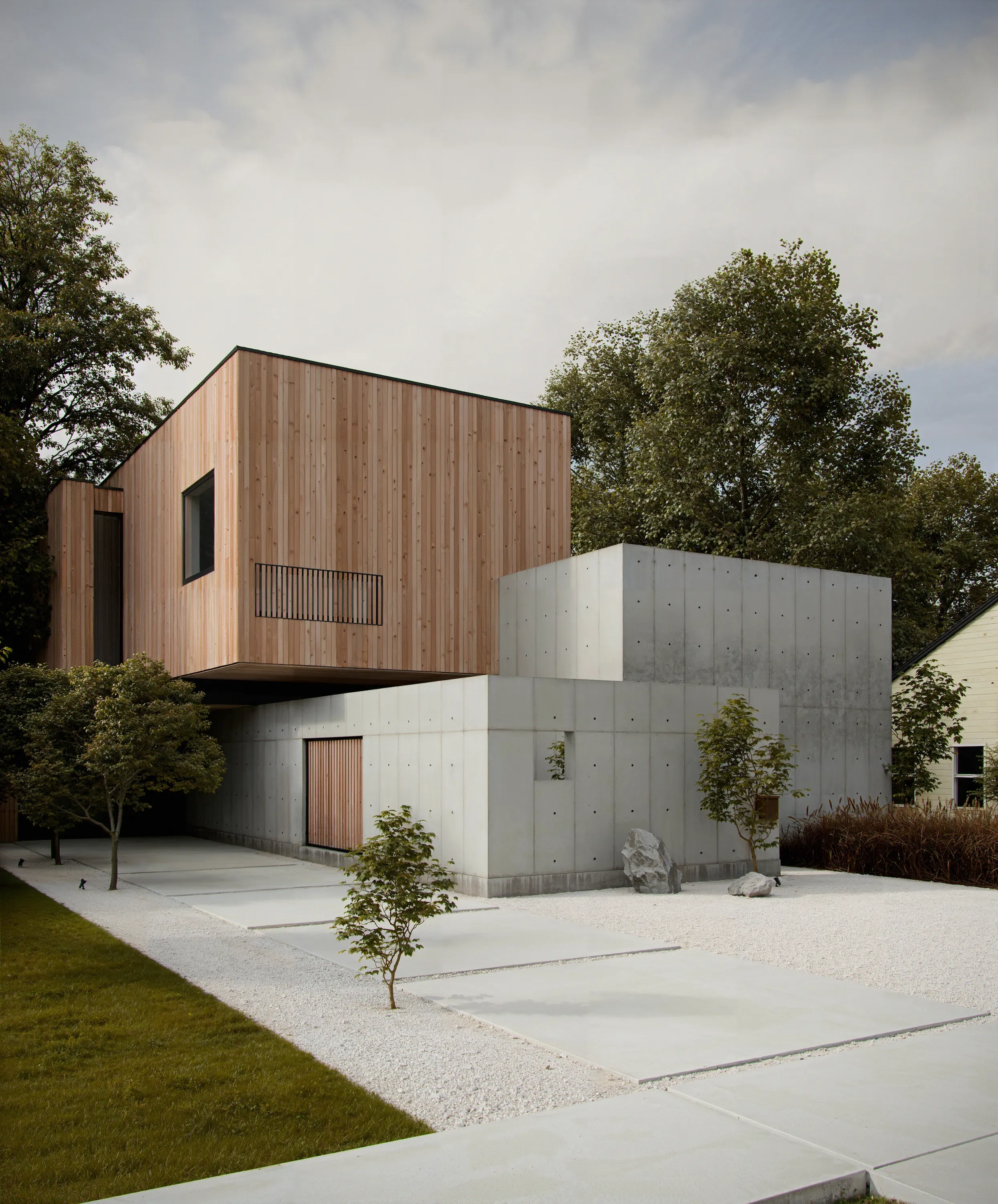 Modern house with wooden upper level and concrete lower level, surrounded by minimalist landscaping with small trees, gravel, and concrete slabs.