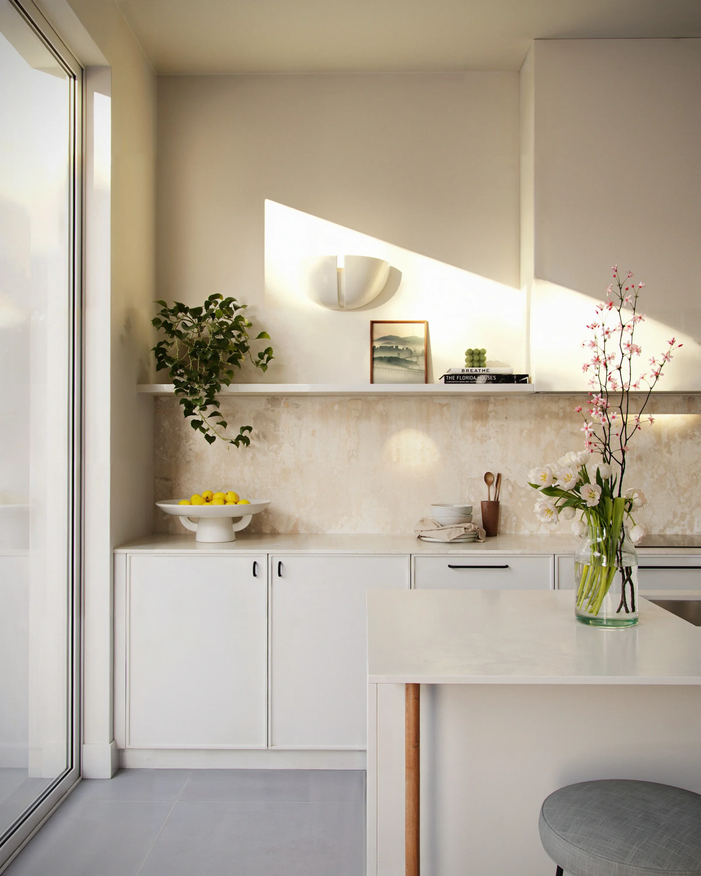 A modern kitchen with white cabinets, a white countertop, and decor including a potted plant, a fruit bowl with lemons, and a vase of pink and white flowers. Sunlight streams in, casting shadows on the wall.