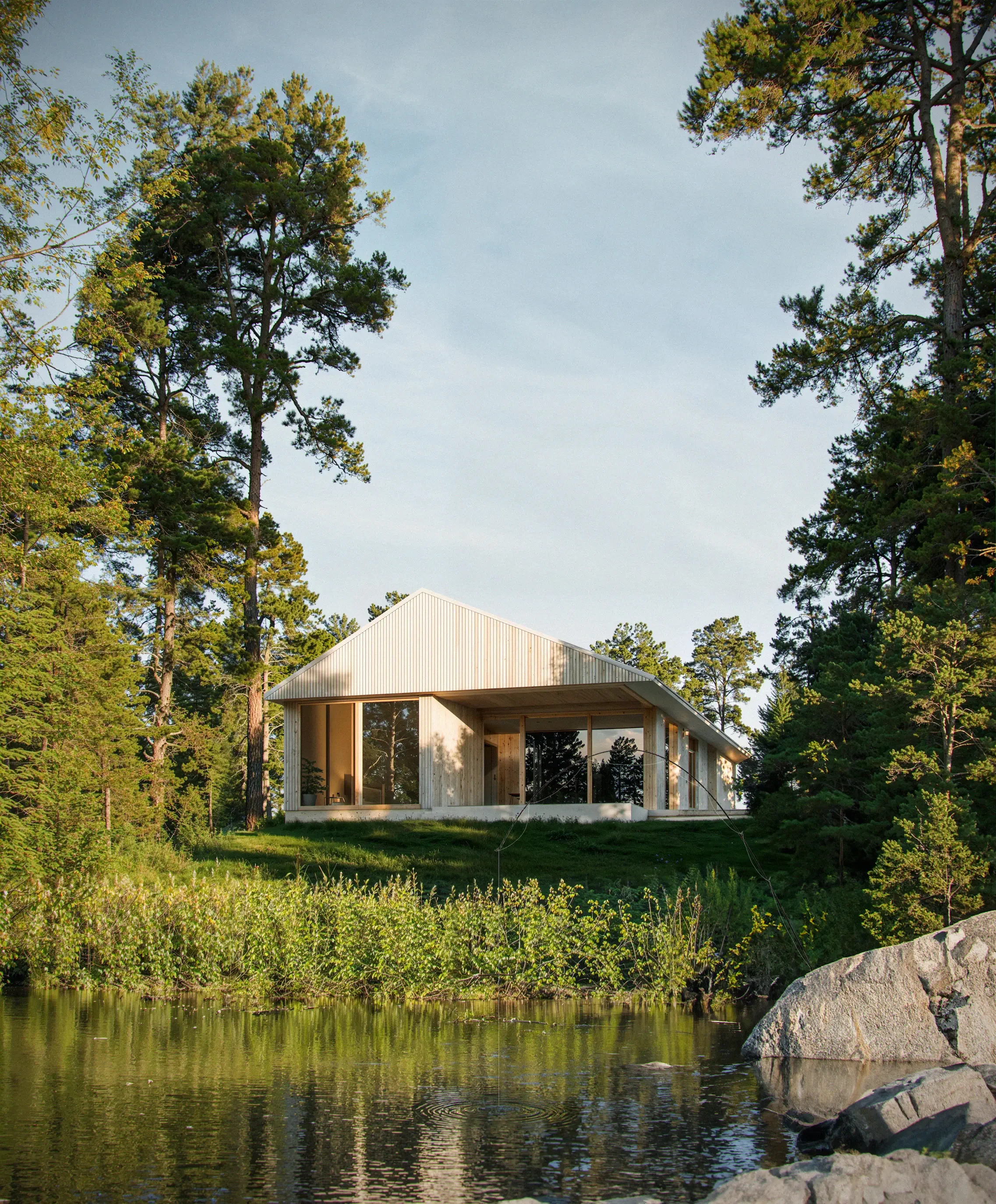 Modern house with large windows on a grassy hill beside a body of water, surrounded by tall trees and rocks, under a clear sky