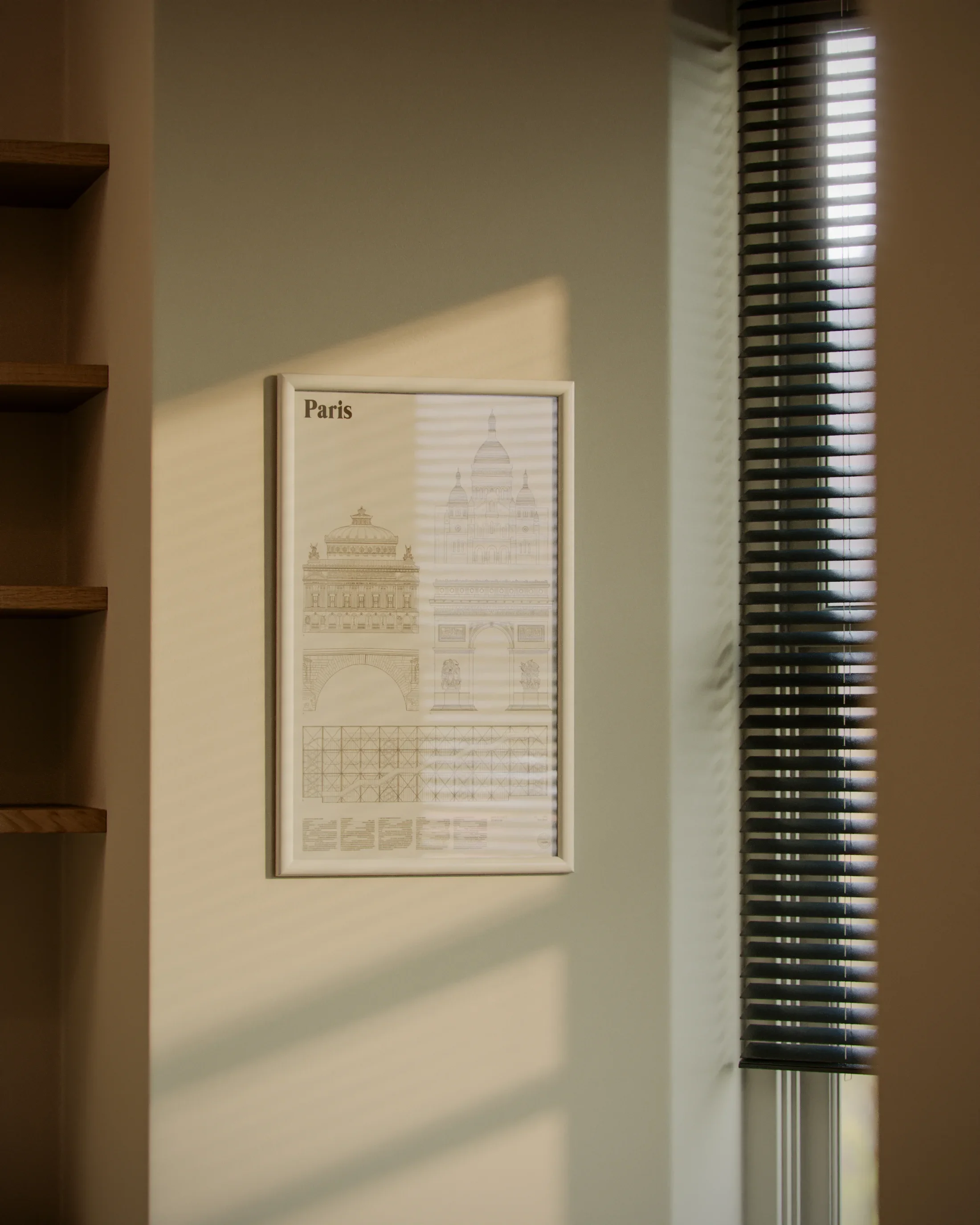 A framed print of architectural sketches of Paris buildings hanging on a light green wall, with sunlight casting shadows through closed blinds.
