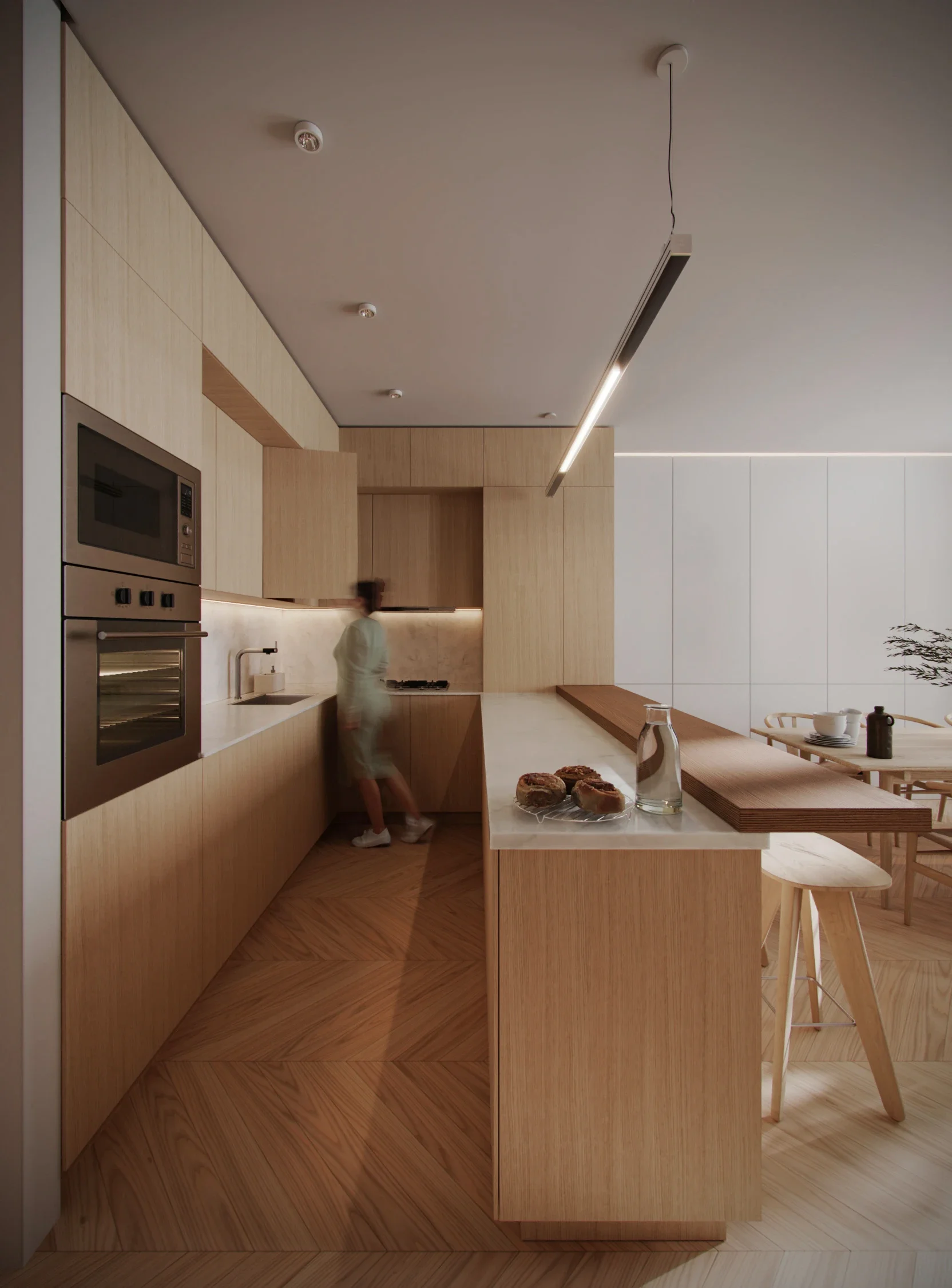 Modern kitchen with wooden cabinetry, a white countertop, and built-in appliances. A person in white is walking near the sink. There are baked goods and a glass of water on the counter, and a dining table with chairs in the background.