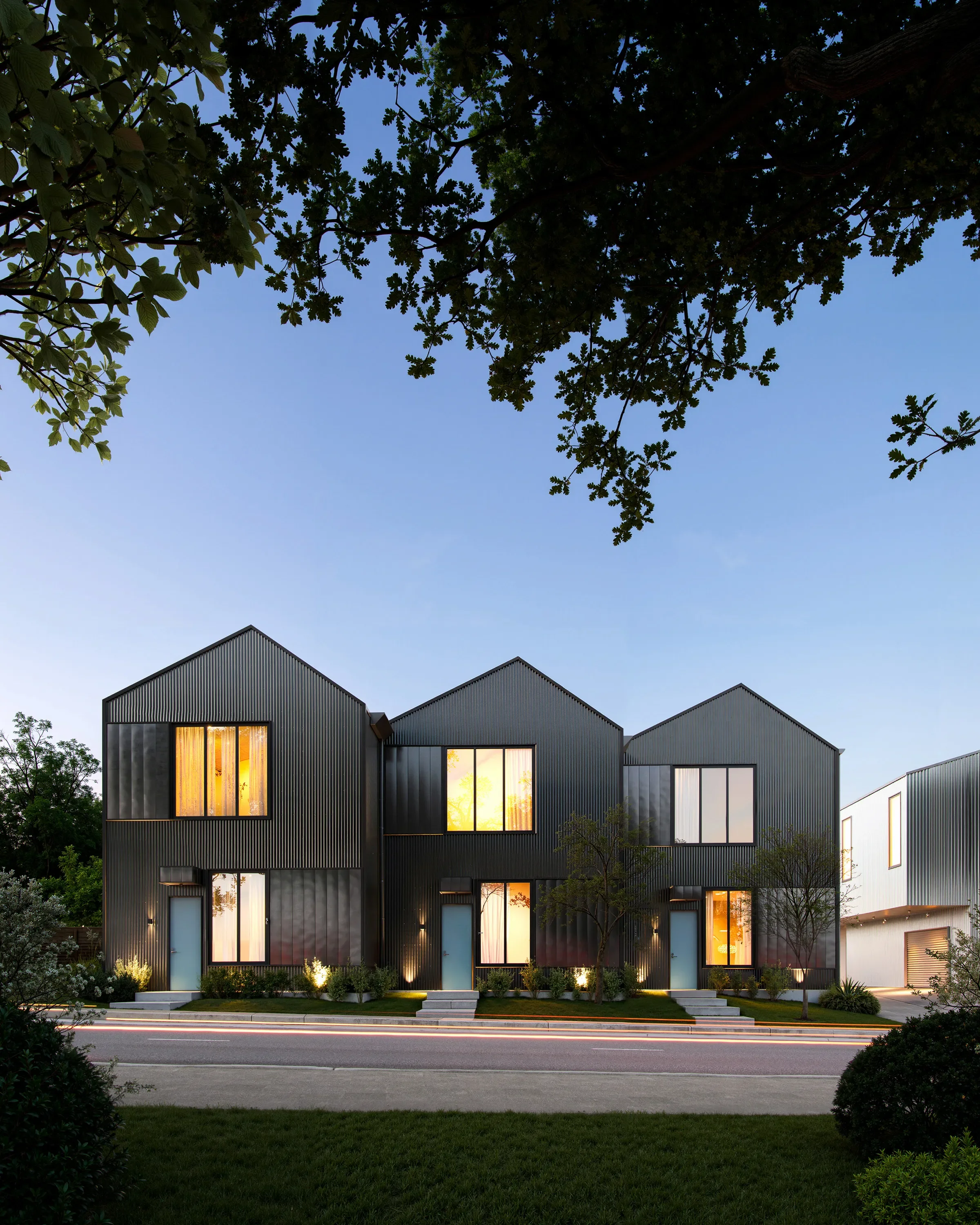 Modern townhouses with black metal siding, lit from inside, in a suburban neighborhood during dusk, with trees and a street in the foreground.