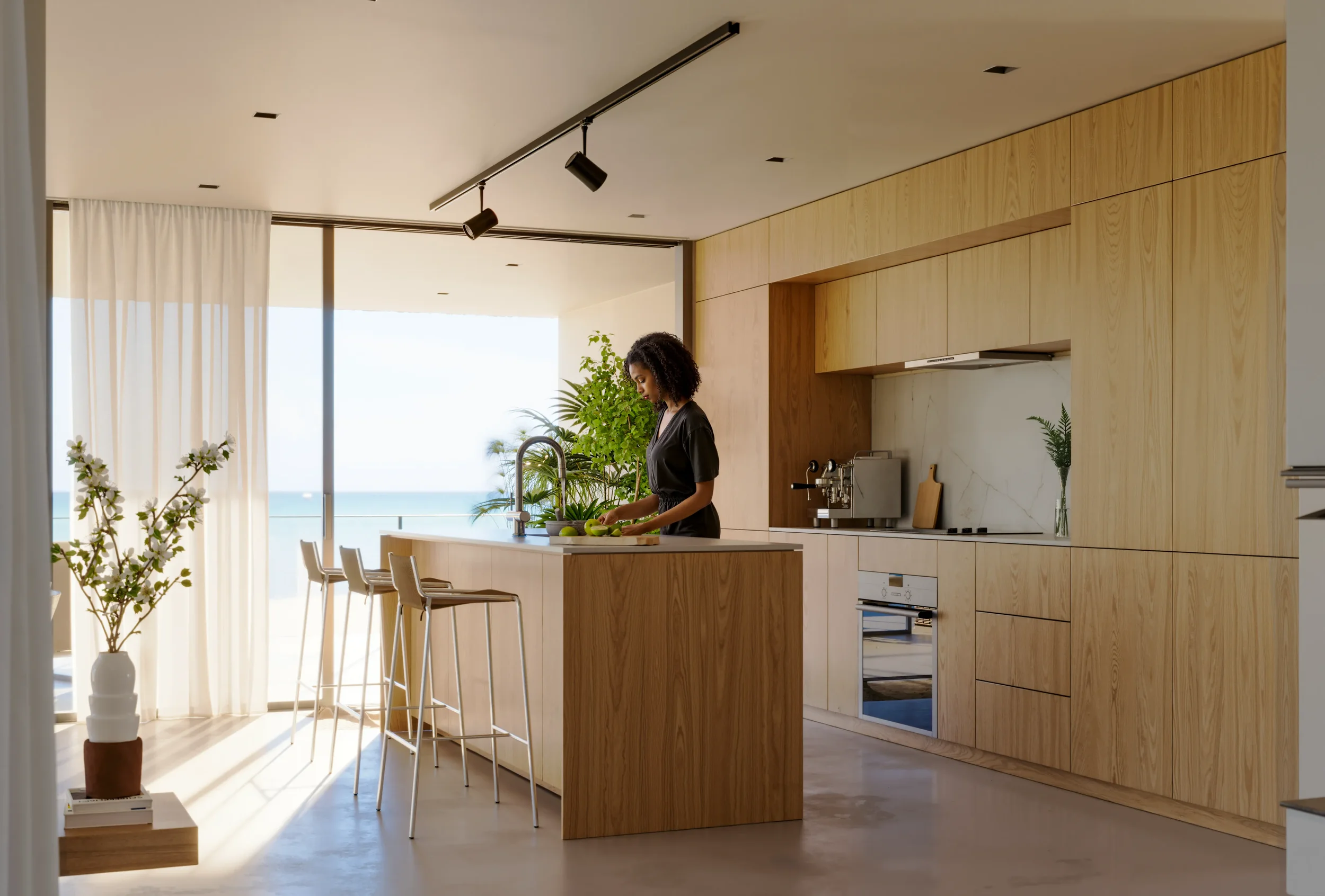 A woman in a black shirt preparing food at a kitchen island with a view of the ocean outside the large glass doors, inside a modern wooden kitchen with plants and minimal decor.
