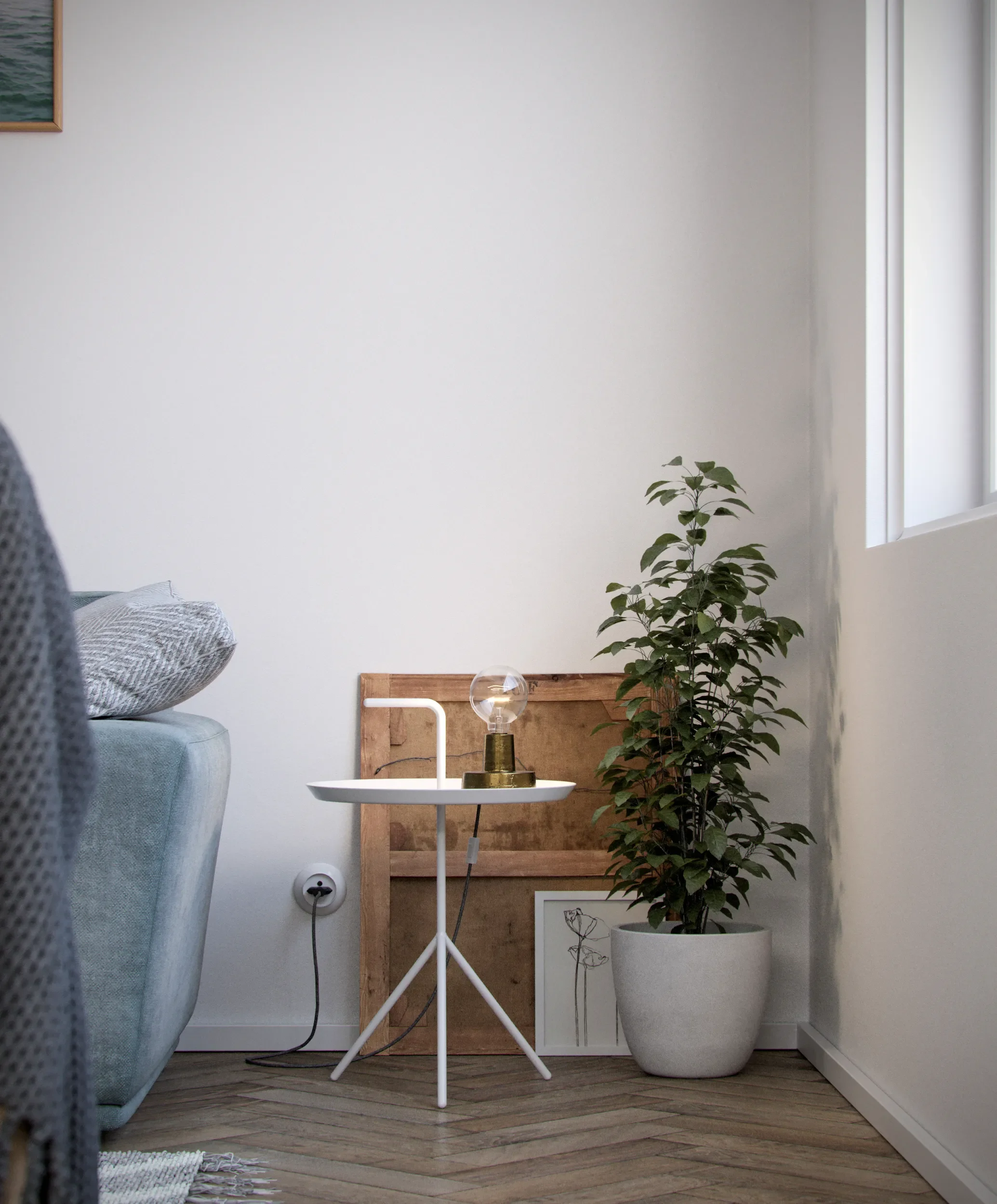 A corner of a living room with a potted plant, a small white side table, a lamp, framed art on the floor, and a plush sofa with cushions.