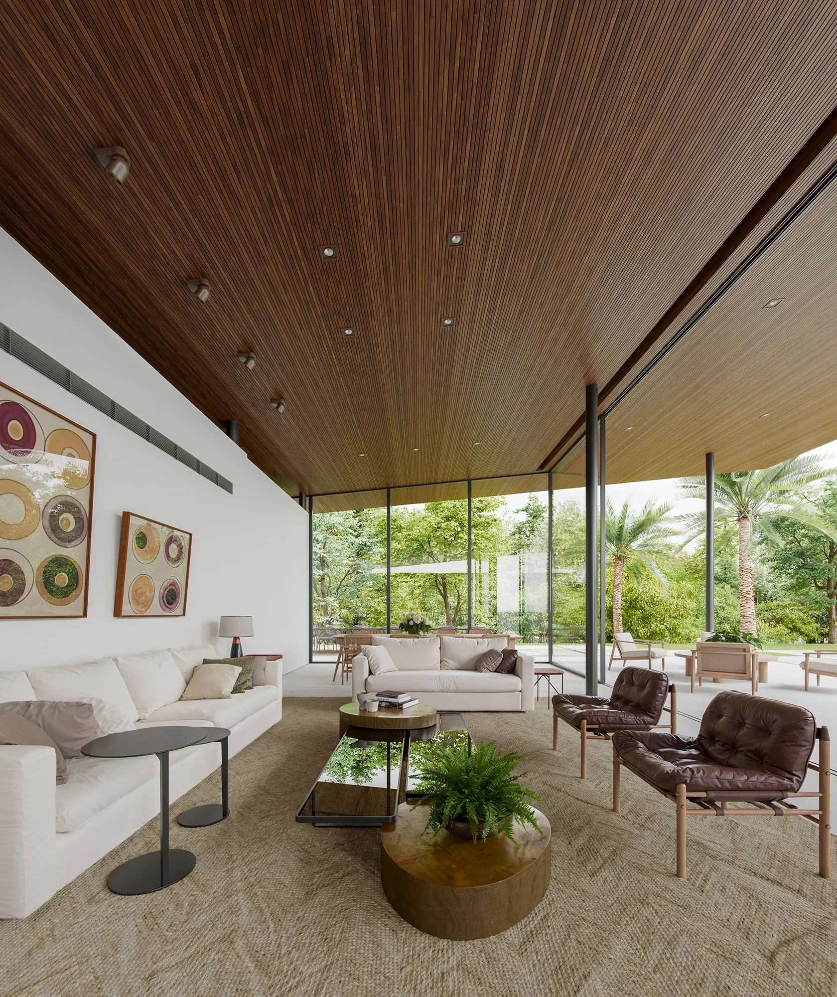 Modern living room with white sofas, leather chairs, and glass windows overlooking a green outdoor area with trees and palm trees.