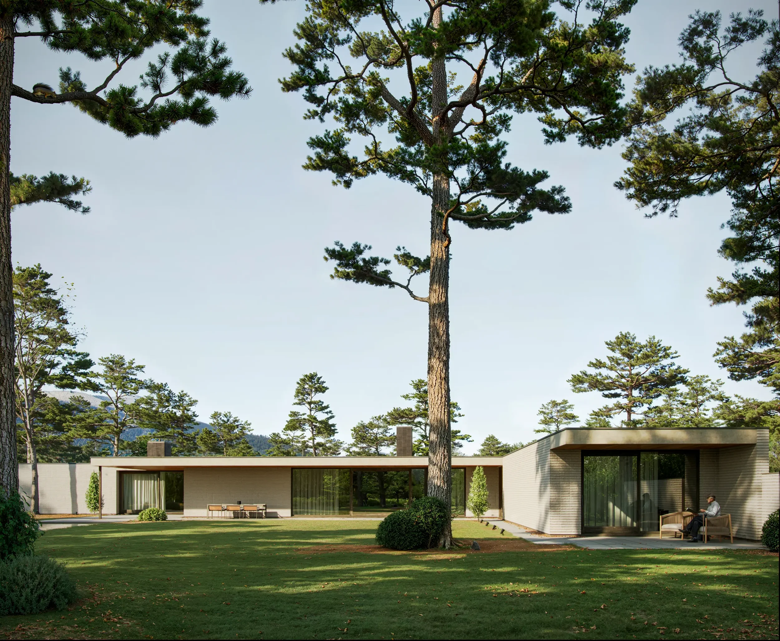 Modern single-story house with large glass sliding doors, surrounded by tall pine trees, with a well-maintained lawn and a person sitting on a bench outside.