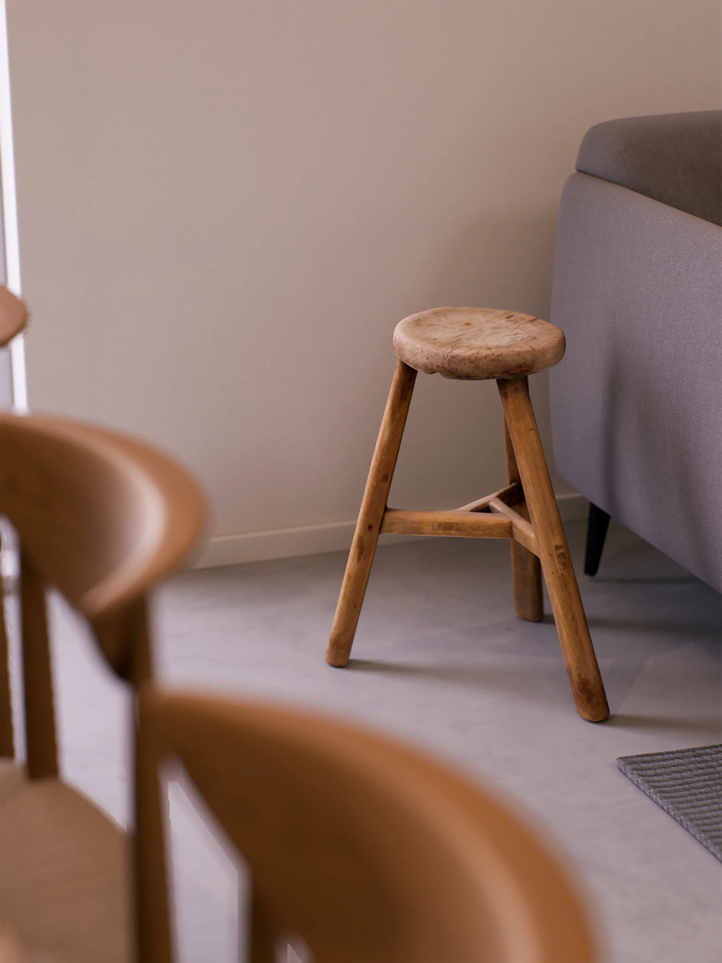 A wooden stool with three legs next to a gray upholstered sofa in a room with light-colored walls and flooring.