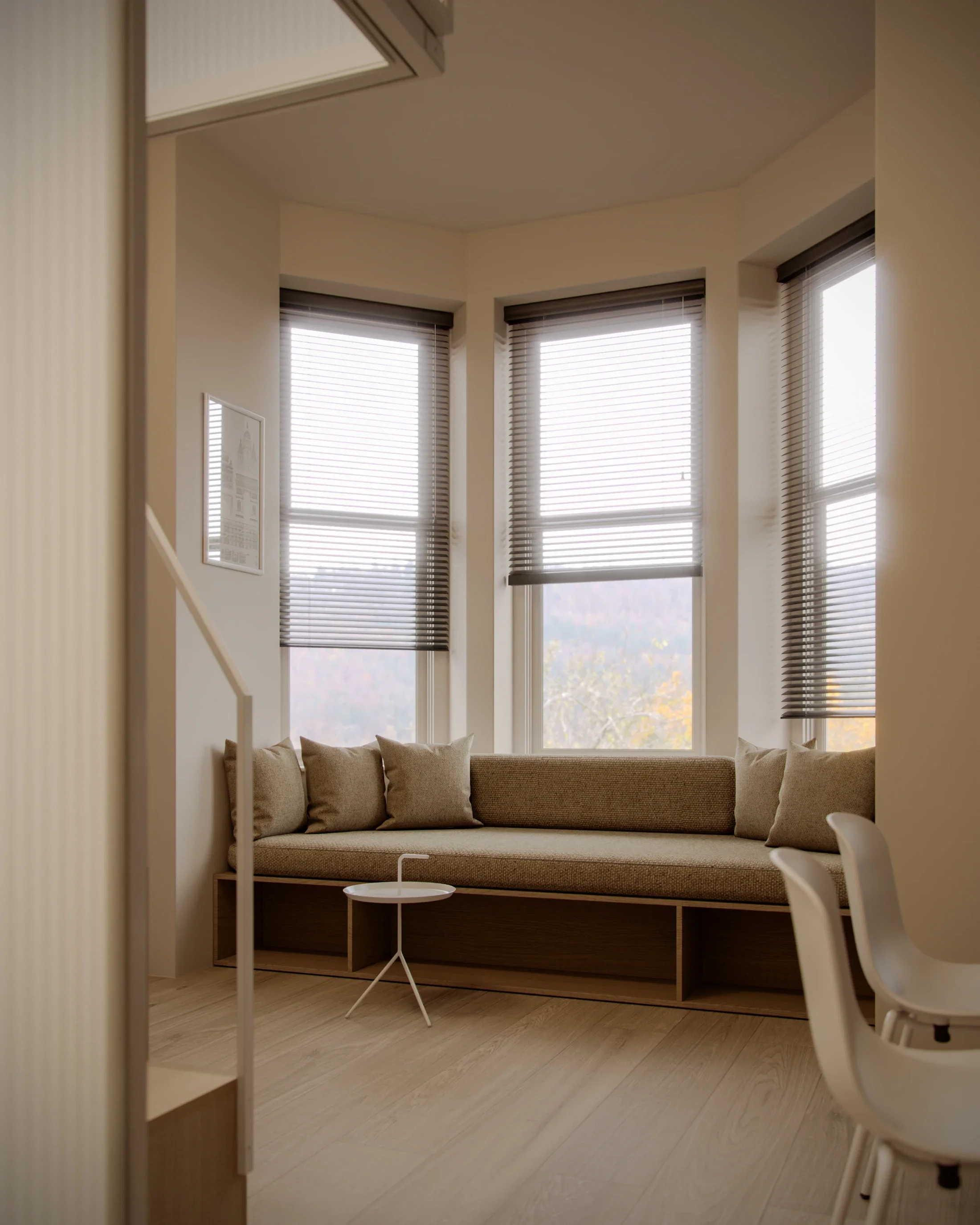 Interior of a cozy corner with large windows, beige cushioned built-in seating, beige pillows, a small round white side table, a white chair, and beige wooden flooring.