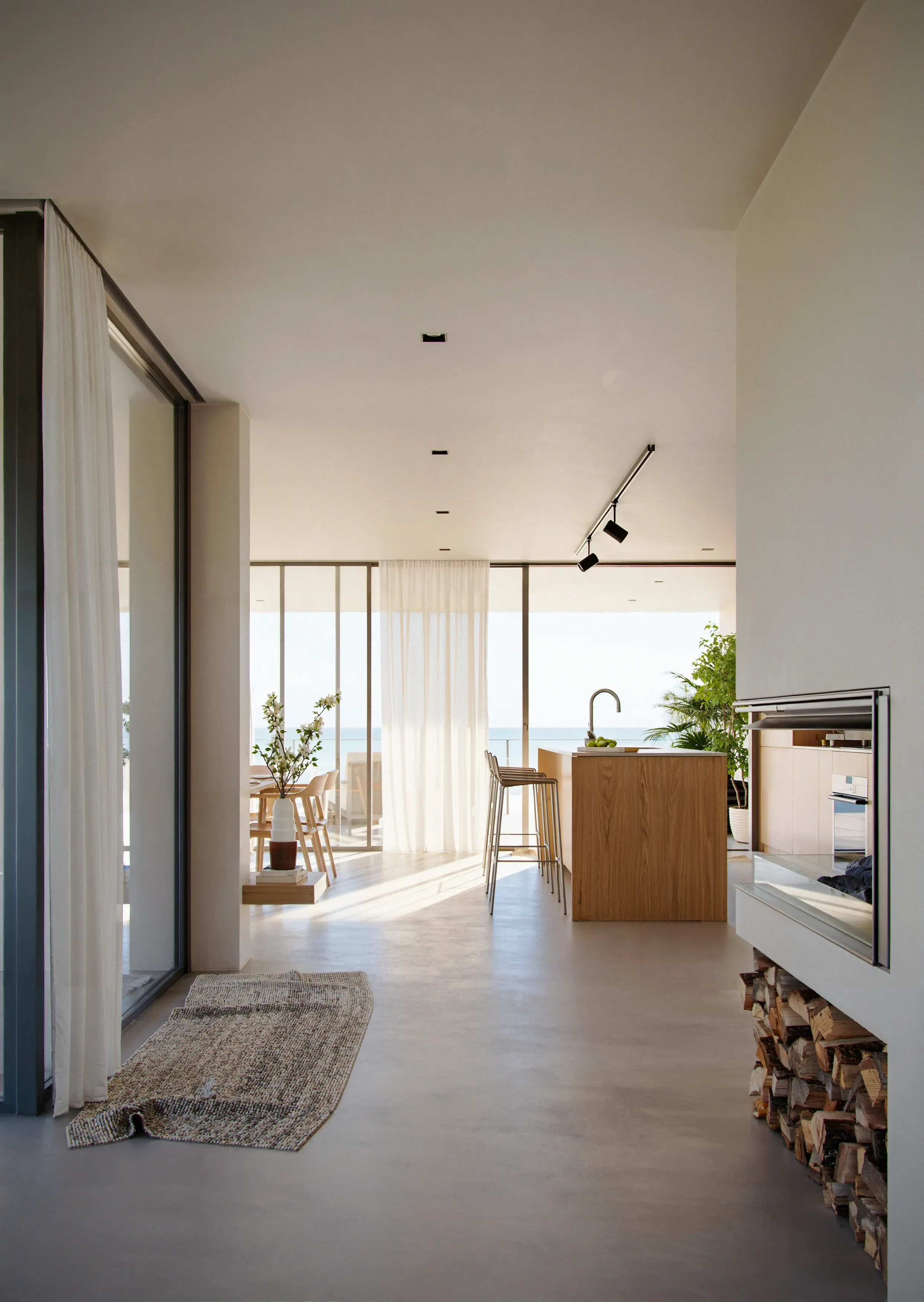 Modern open-concept living room and kitchen with large windows overlooking the ocean, wooden furniture, a potted plant, and a stack of firewood.