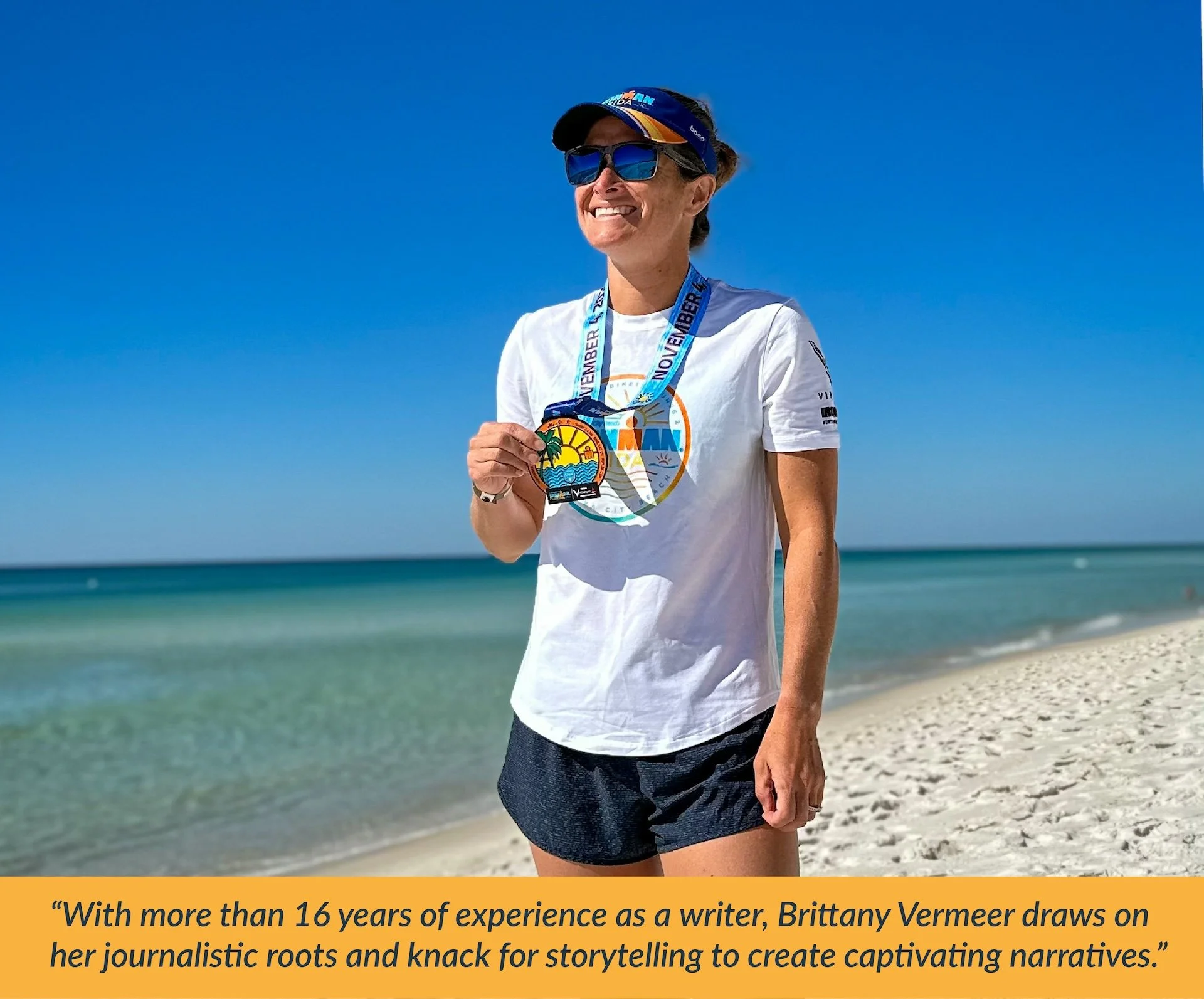 Female triathlete  standing on the beach holding an Ironman medal, wearing sunglasses and a cap, with the ocean and a clear blue sky in the background.