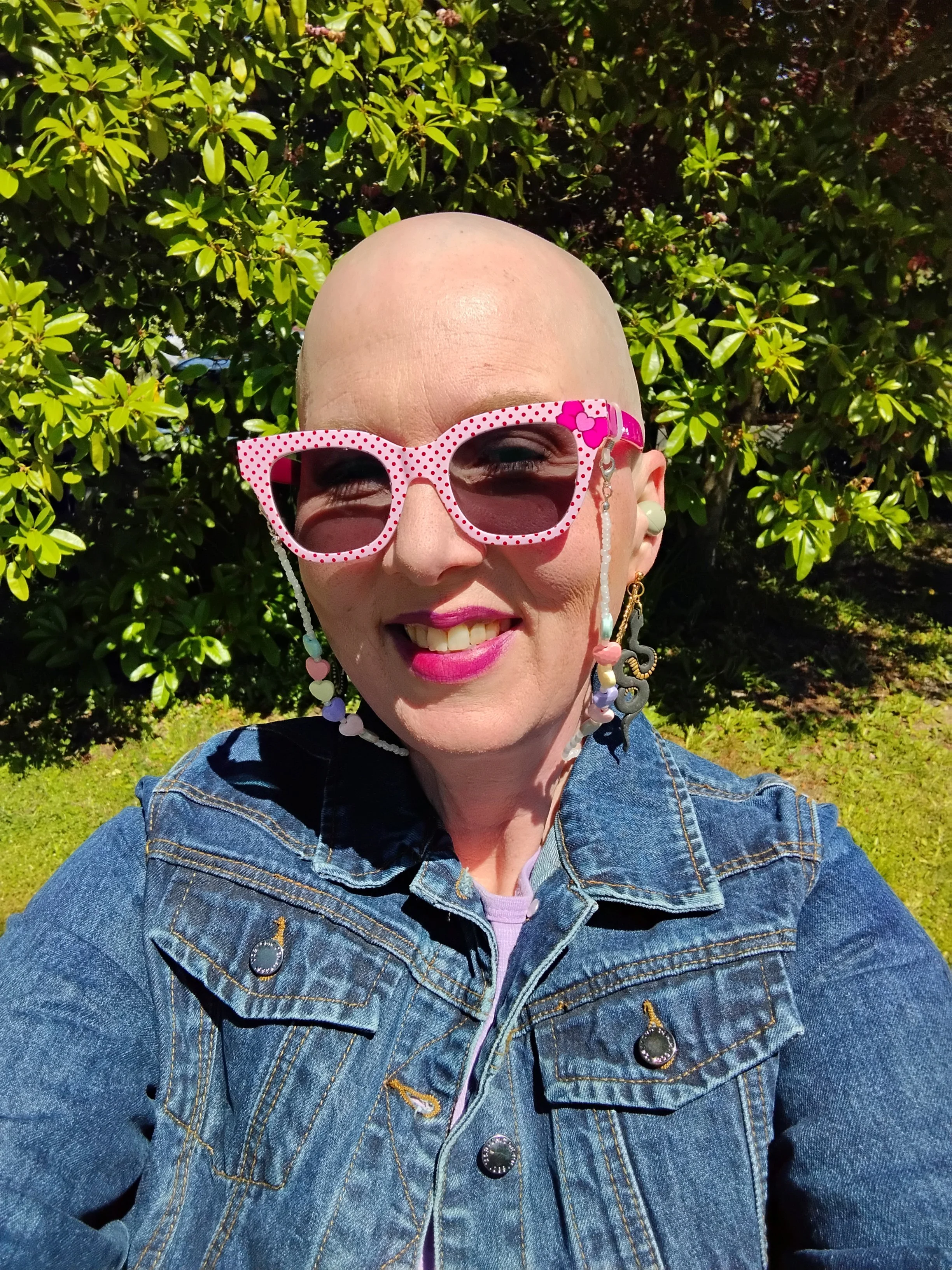 Selfie of a woman with her head shaved wearing a blue denim jacket and glasses with lively pink frames that have polka dots and clowers on them; she is standing outside in front of bushes in the sun