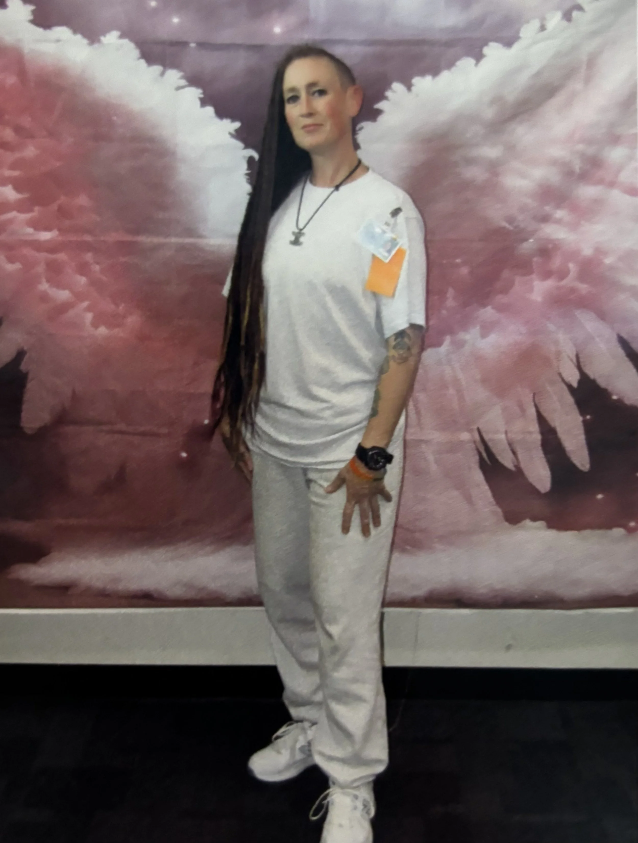 A woman with long dark hair and an undercut standing in front of a background of dark pinks and light purples.