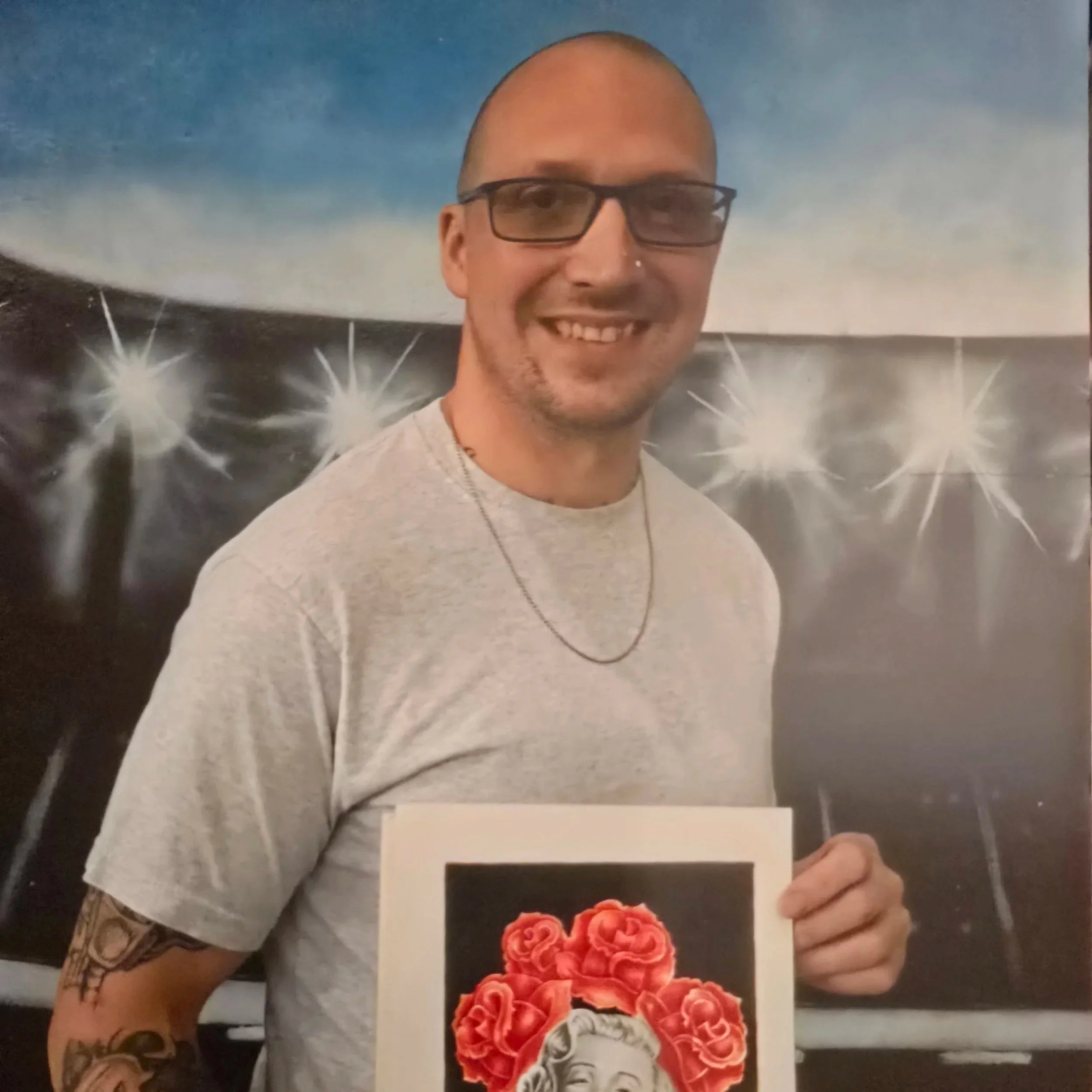 A man with glasses and pale skin wearing a thin necklace and smiling towards the camera holding a drawing of Marilyn Monroe with a crown of red roses.