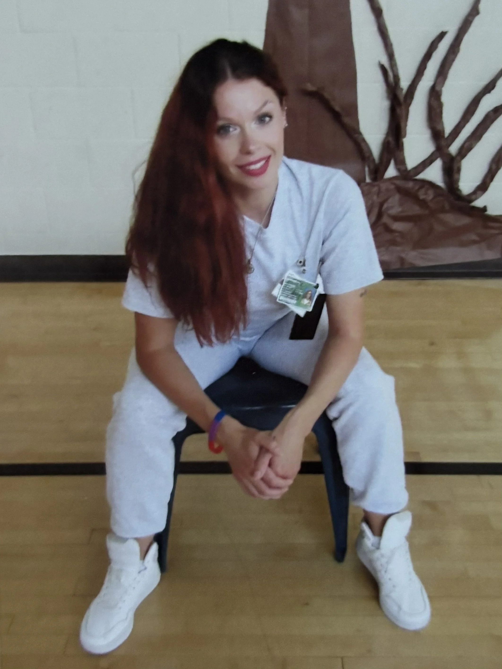 Photo of a woman with dark red hair and all-white clothing sitting on a chair and smiling at the camera.