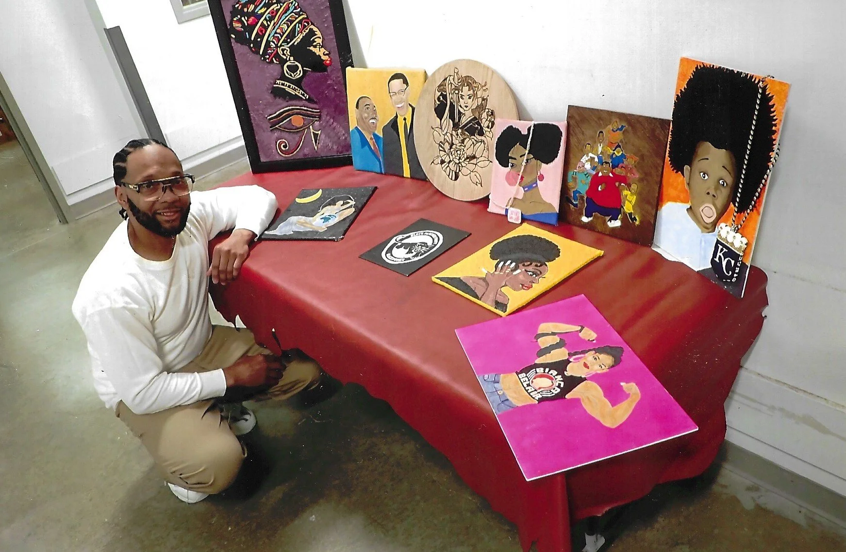 A man kneeling by a table that is featuring his artwork.