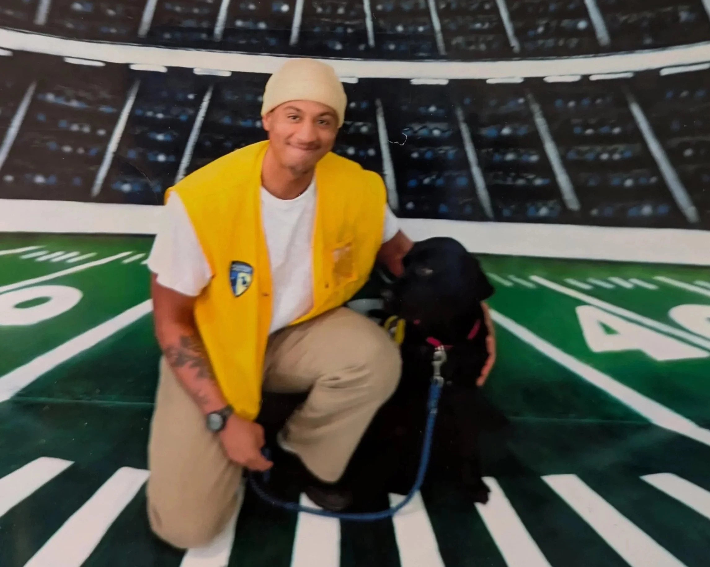 A man with medium brown skin kneeling with his arm around a black dog and smiling.