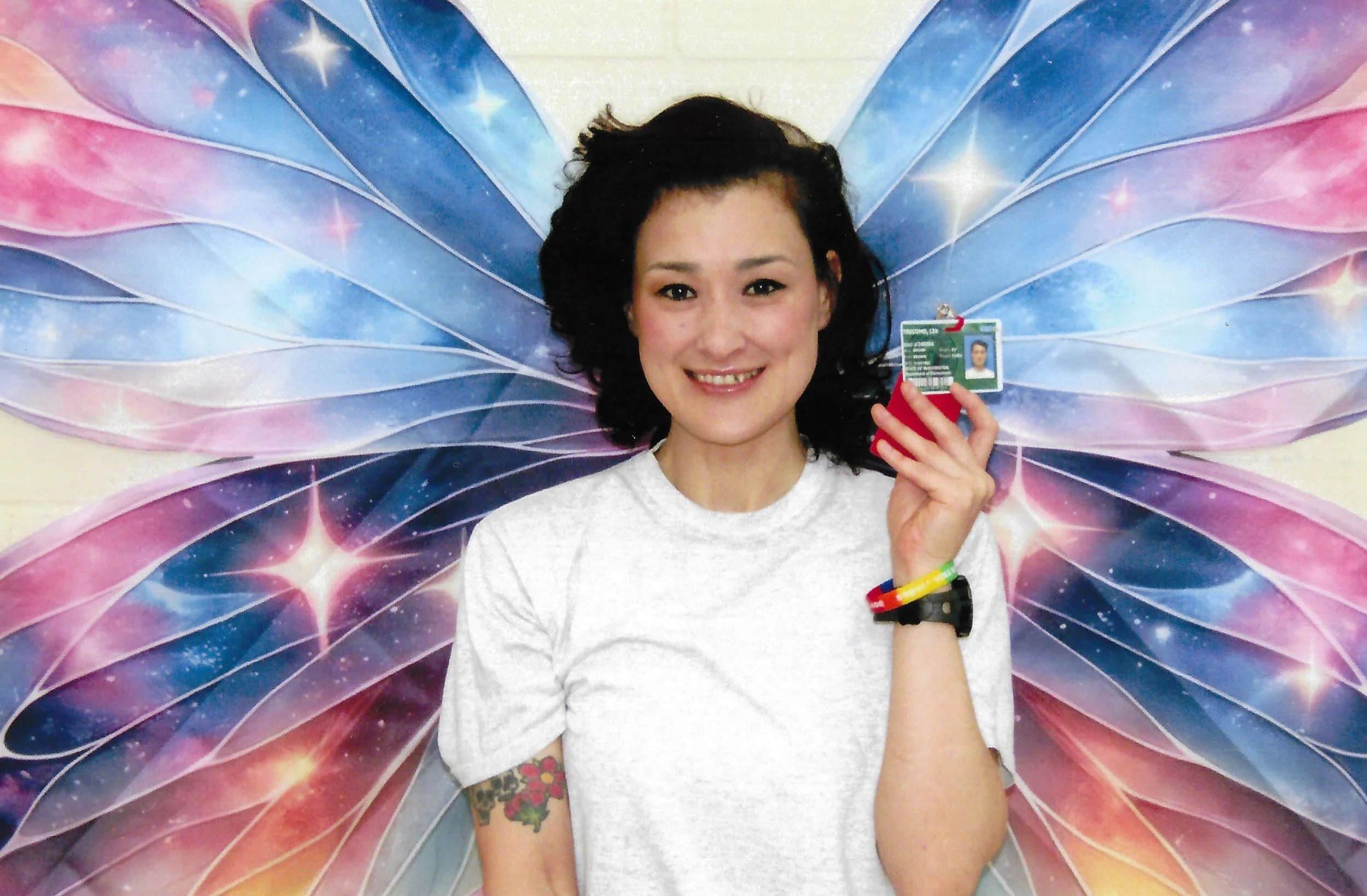 Selfie of a middle-aged woman with brown and red hair hair of mixed Asian descent, framed center from the waist up in a white prison uniform t-shirt. She is flanked by two colorful butterfuly wings of cerulean blue and magenta.