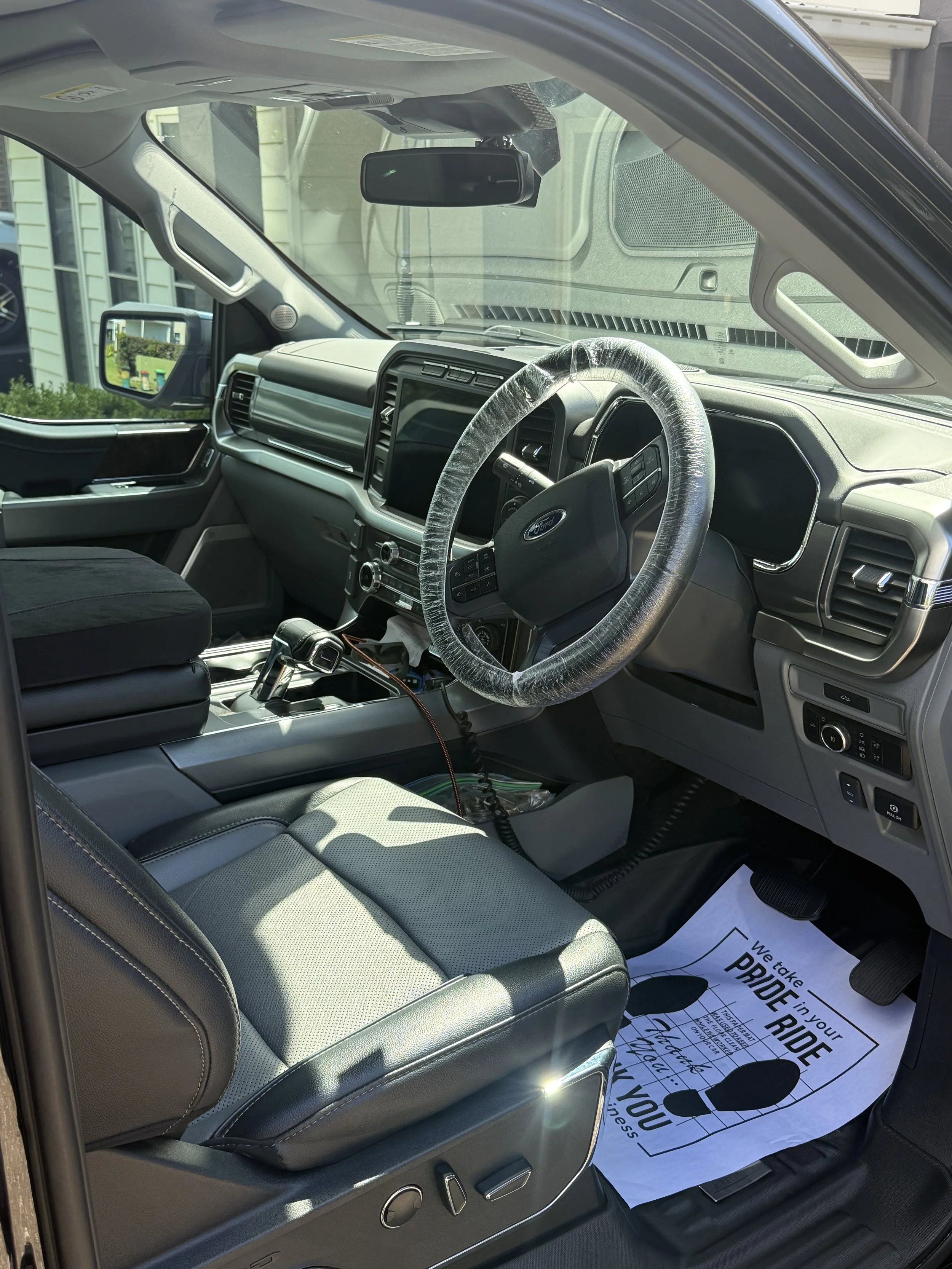 Interior of a Ford vehicle showing the driver's seat, steering wheel with a plastic cover, dashboard, gear shift, and a paper floor mat with an encouraging message.
