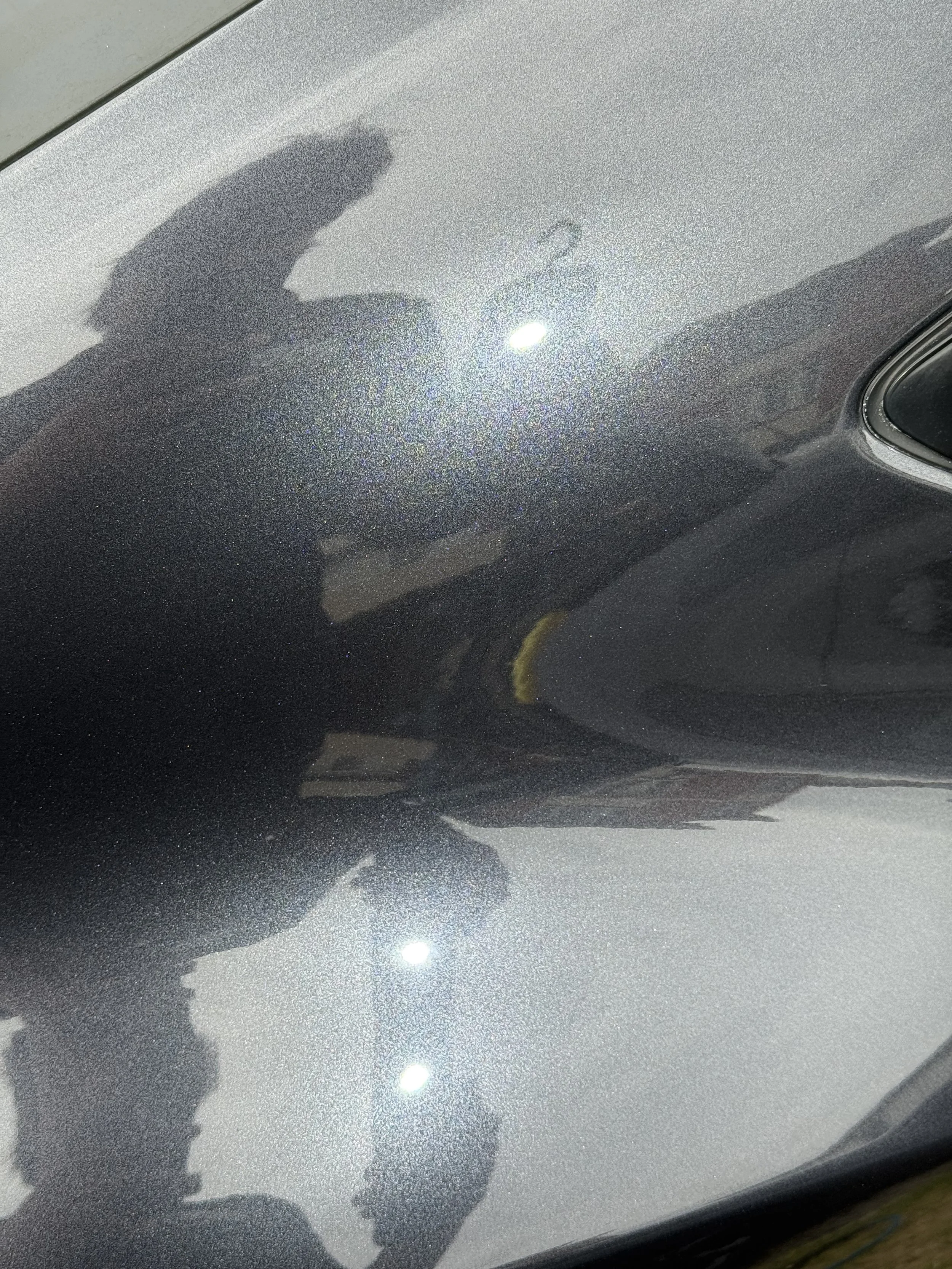 Close-up of a black car's exterior with a metallic finish, showing reflections of the person taking the photo and the bright light sources.