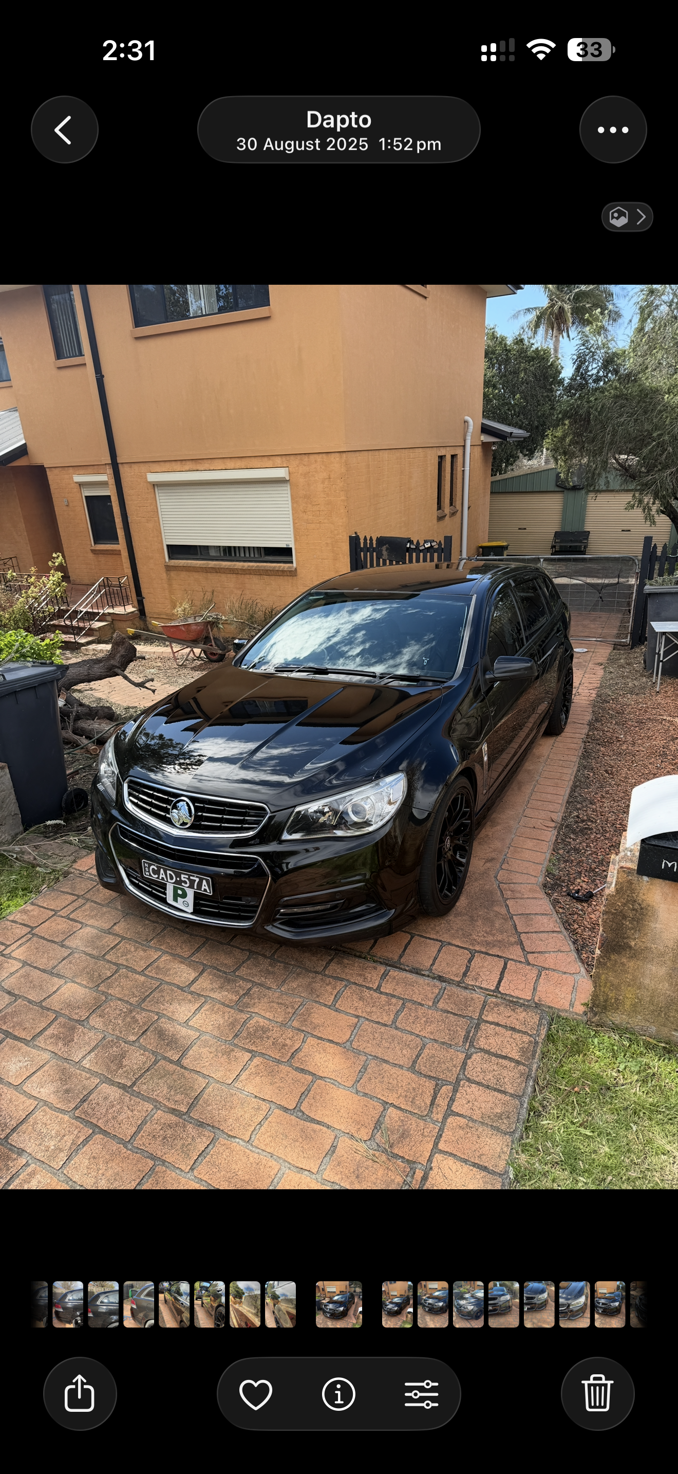 Black sedan car parked on a brick driveway outside a tan-colored house with windows, a backyard with trees, gardening tools, and a fence.