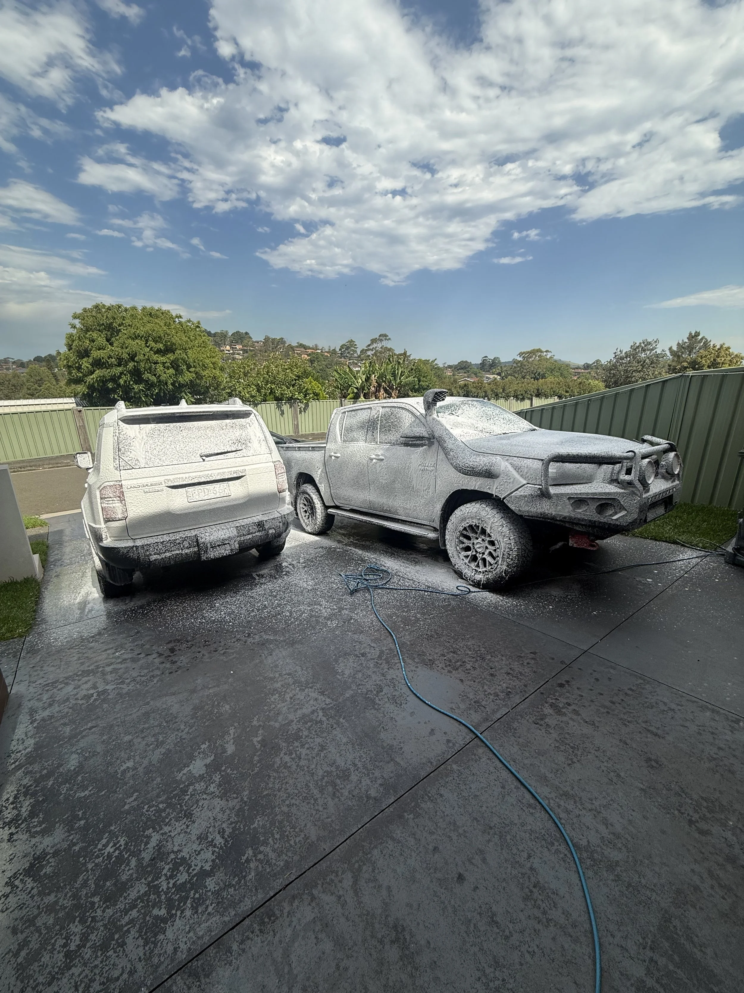 Two cars being washed with foam on a driveway under a partly cloudy sky. One car is covered in thick foam, and the other is being washed with foam on it.