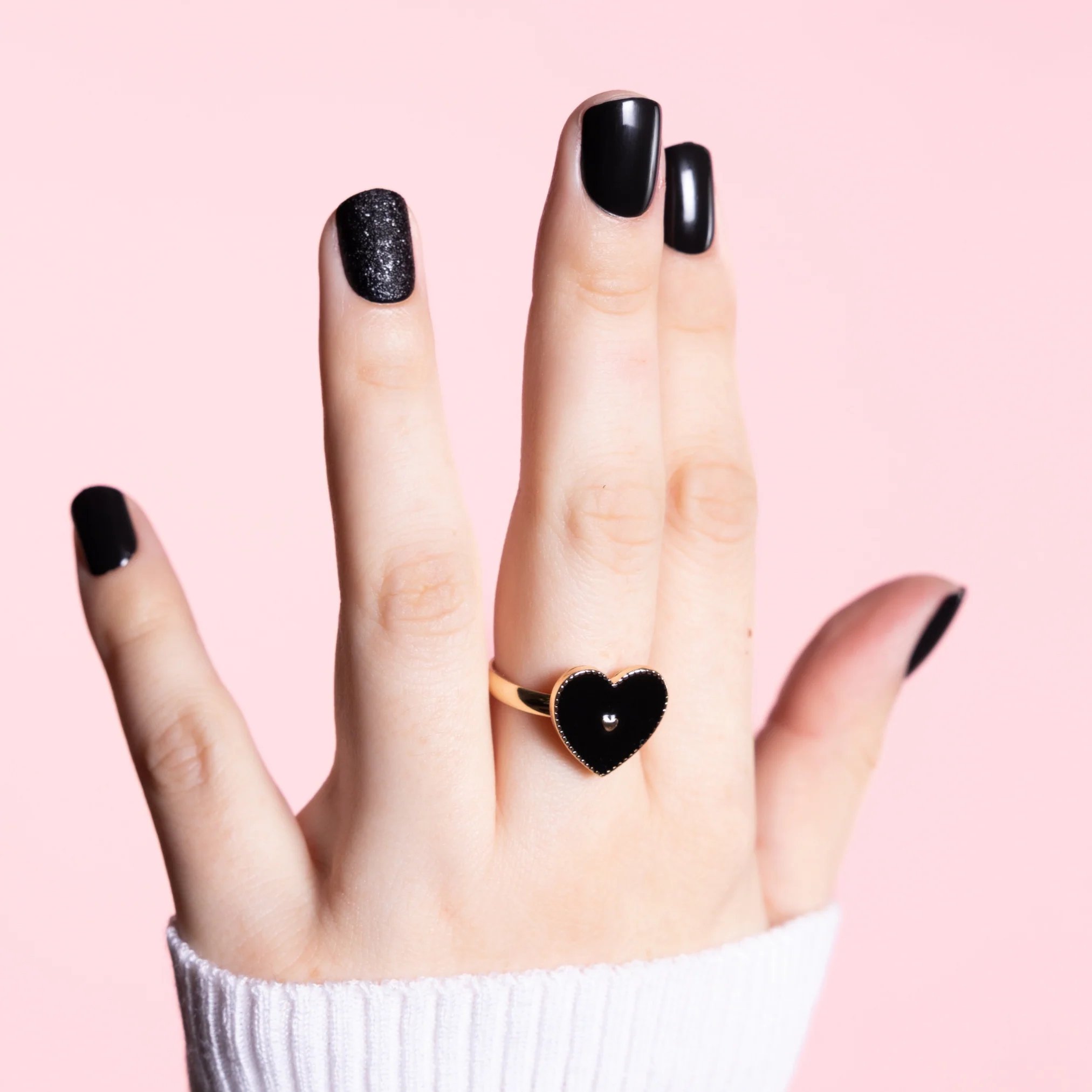 Hand with black manicured nails wearing a gold ring with a black heart design on a pink background.